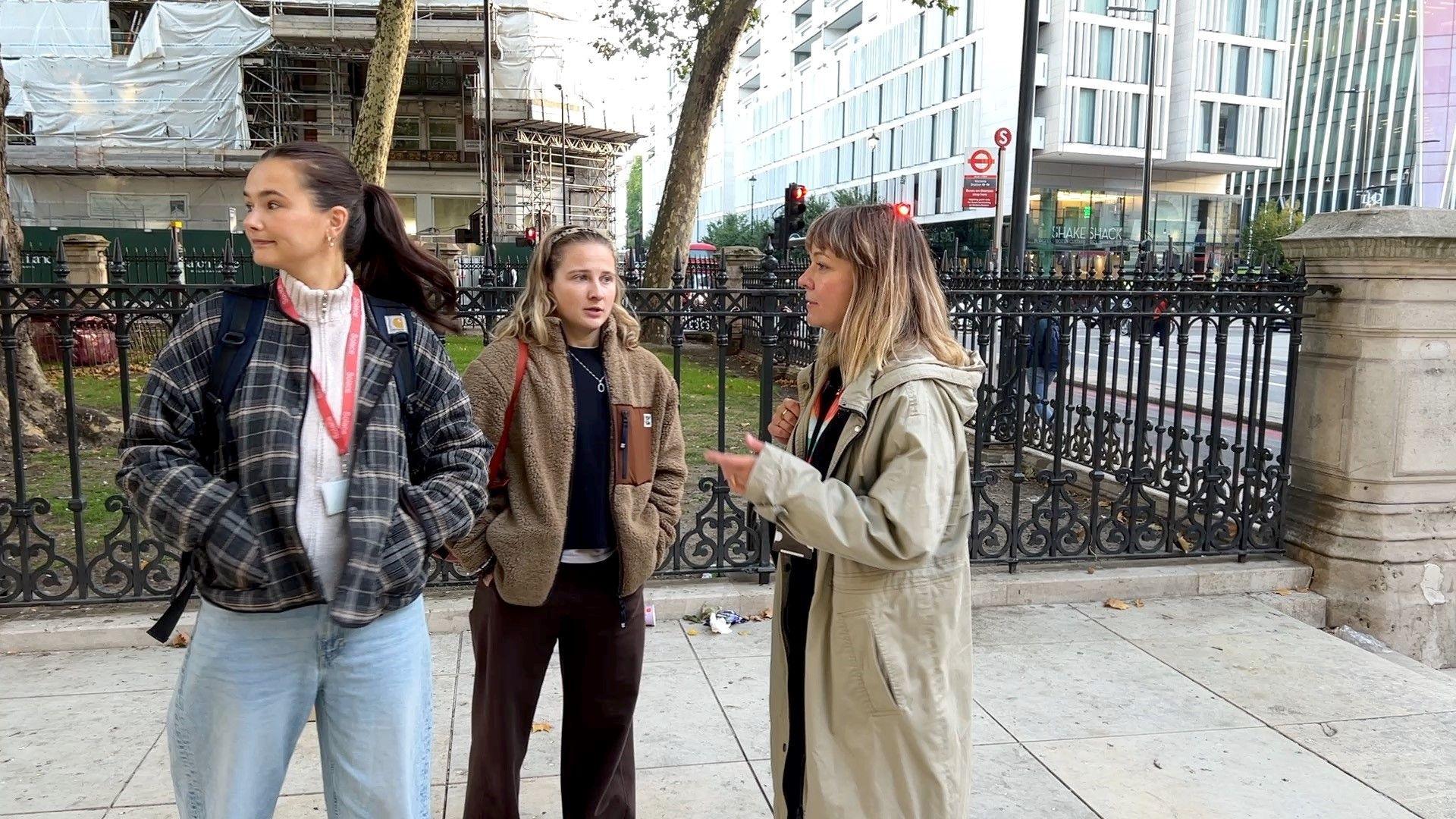 Three young women wearing lanyards have a discussion near a park