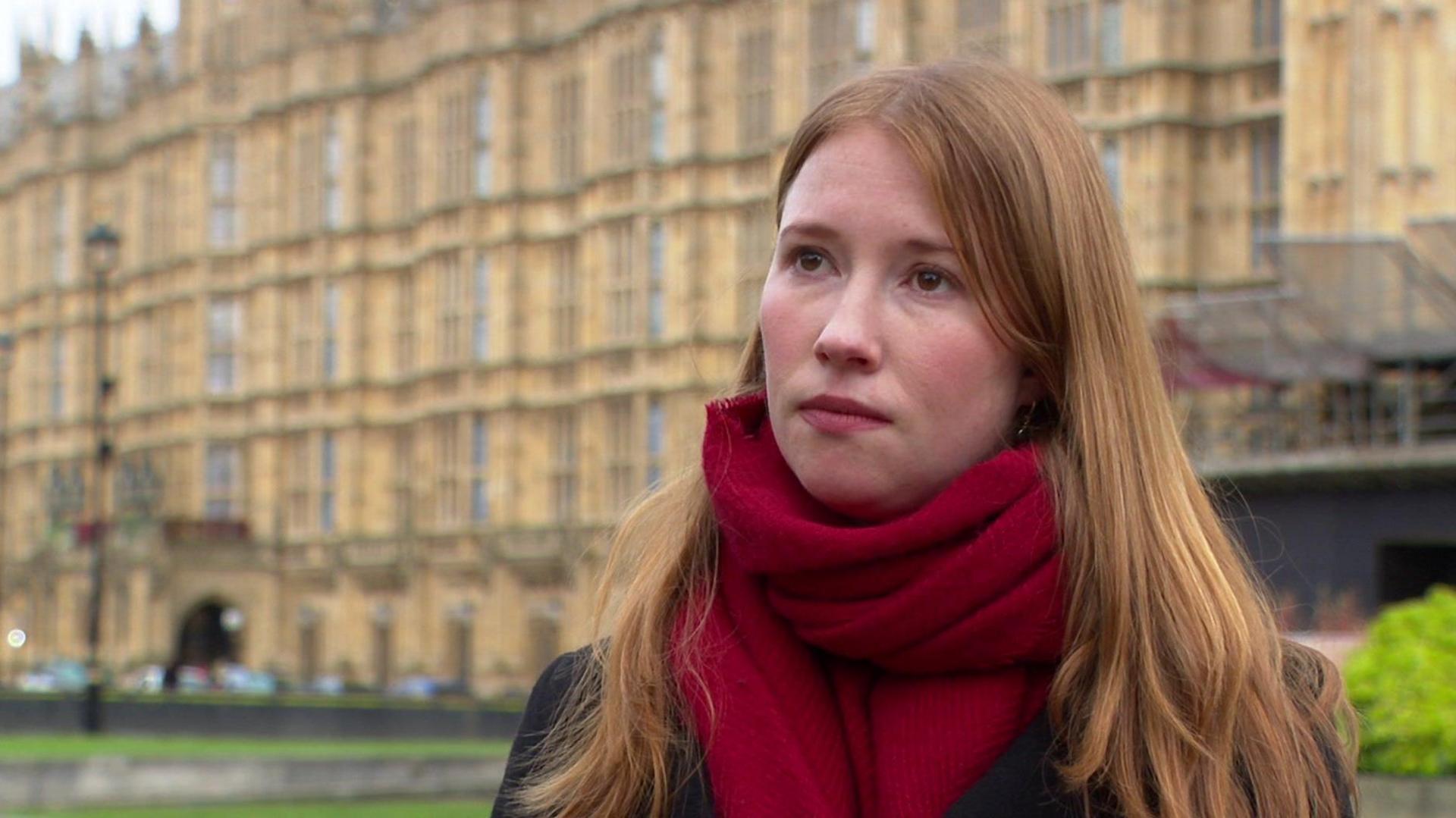 Helena Dollimore stands in front of the Palace of Westminster. She is wearing a dark coat and a red scarf.