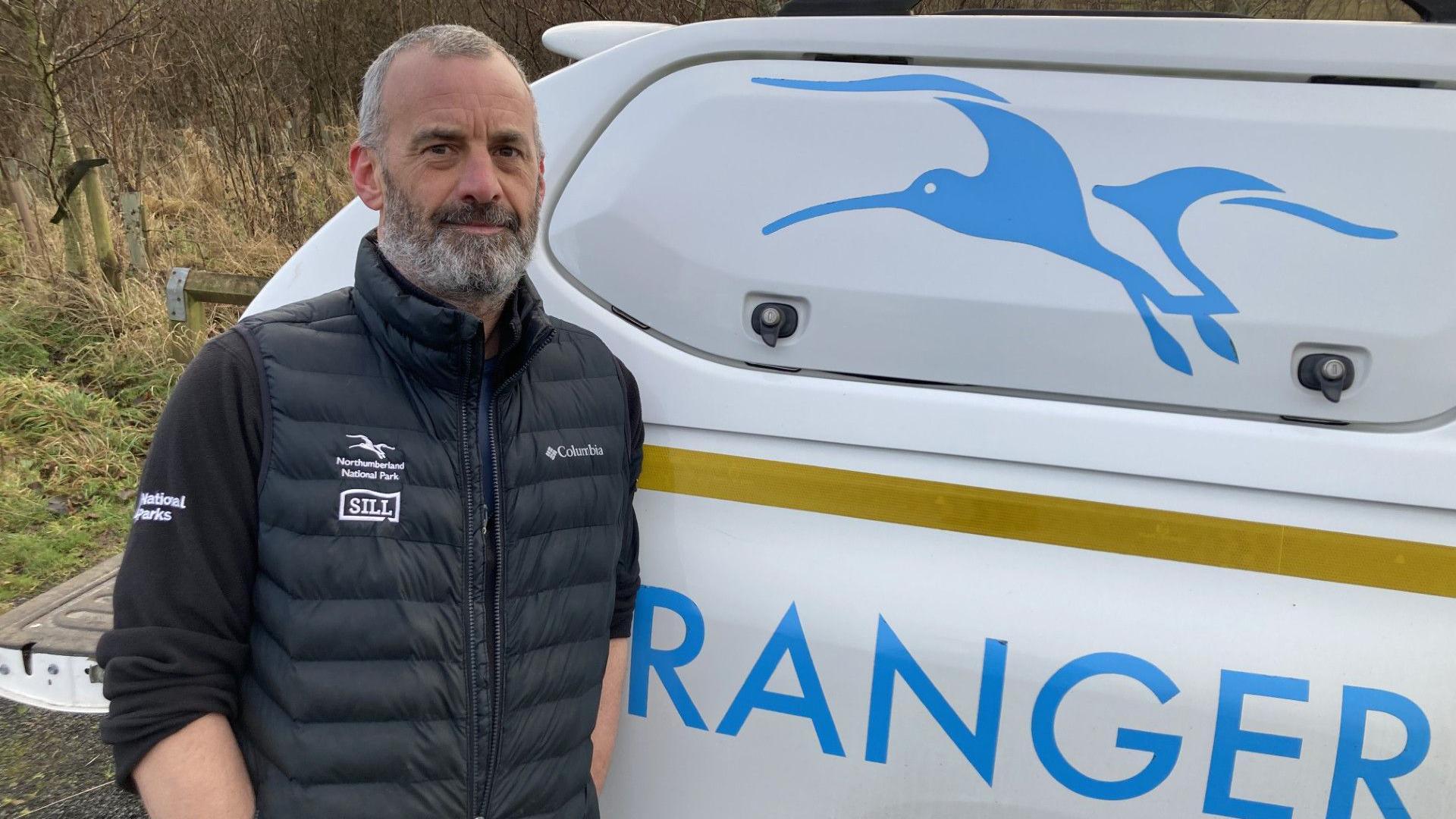 Gary Pickles is standing next to a van with the word ranger on it. He is wearing a black jacket with the Northumberland National Park logo on it and has grey hair and a grey beard. He is a man in his 40s or 50s.