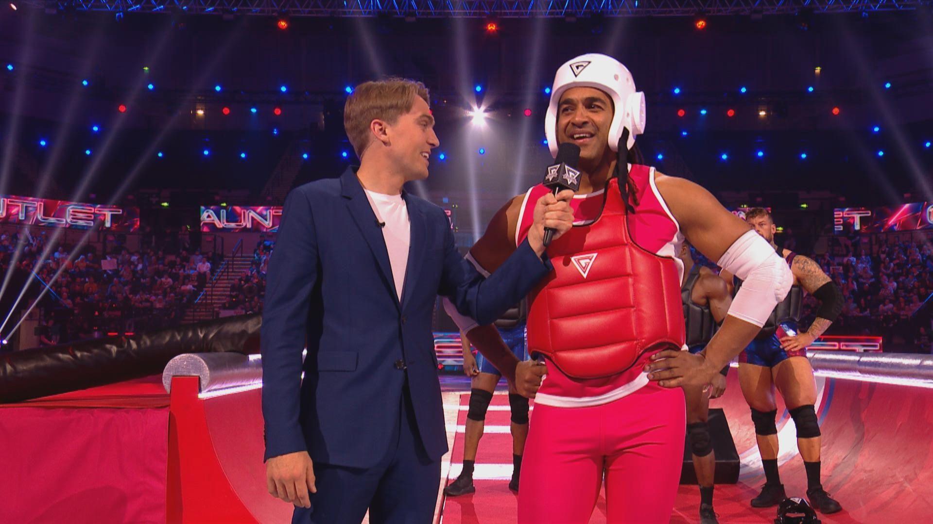 TV presenter Barney Walsh holds a microphone up to Wesley Male, who is clad in red sportswear and wearing protective inflatable jacket and a white helmet. They are on the set of Gladiators.