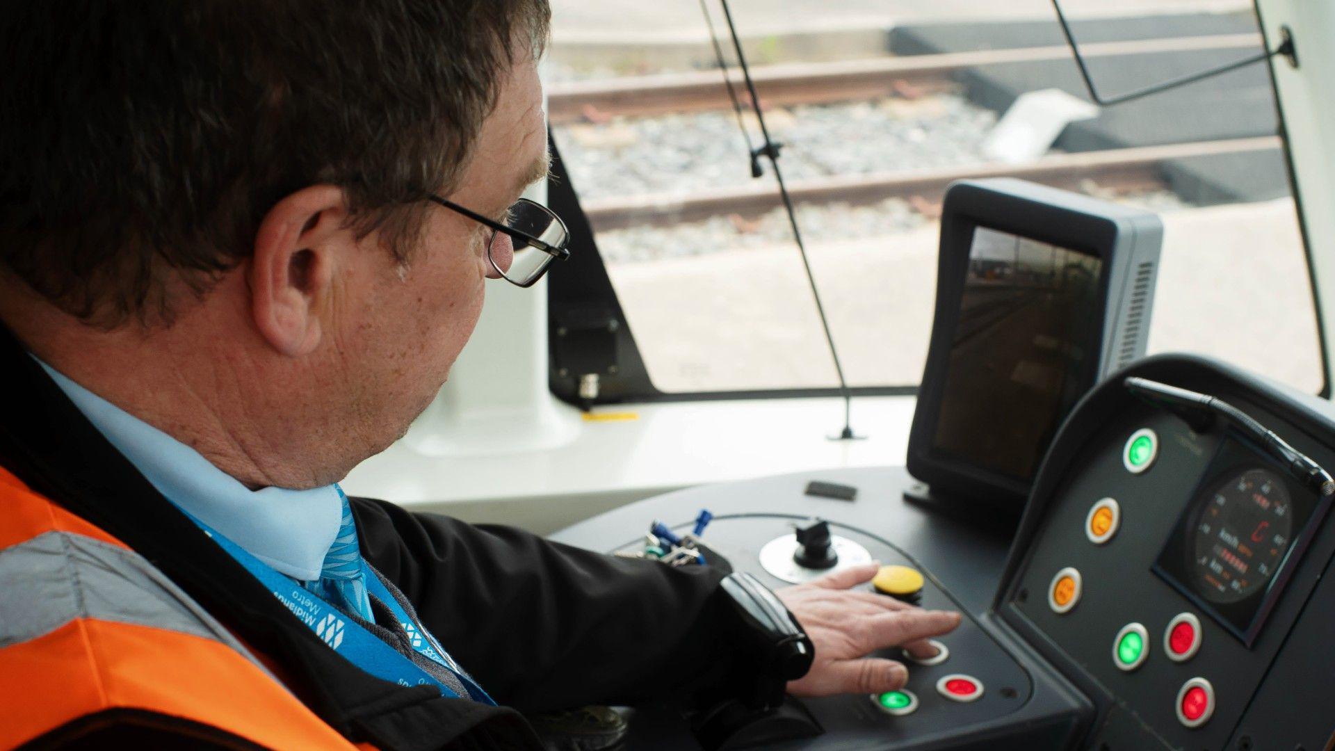 Chris Bruce as the controls of the tram. He has a orange high-vis vest on and is pressing a button on a panel. There are a number of buttons and knobs. 