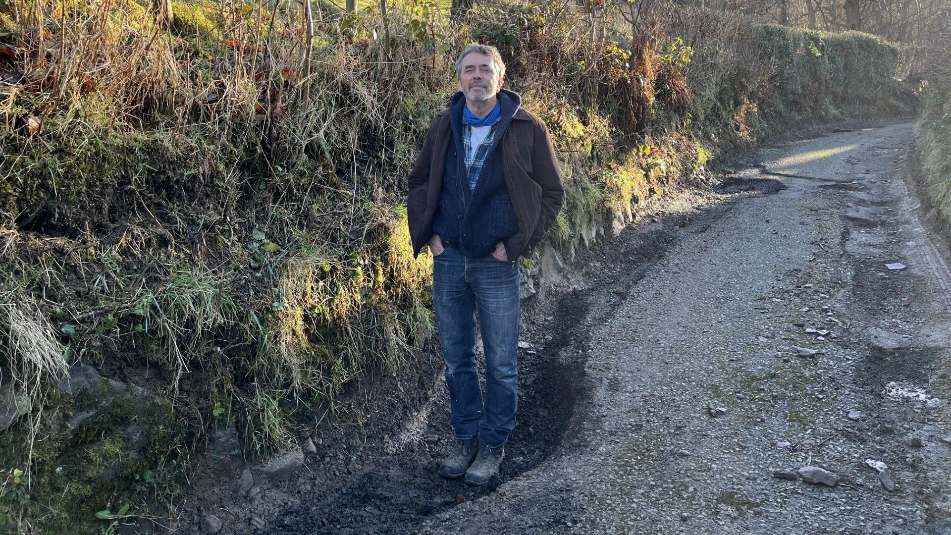 A man with short grey hair and a short grey beard stands in a large pothole on a country lane with his hands in his pockets.  He is wearing a brown jacket over a blue cardigan and a blue and white checked shirt and white t-shirt, blue jeans and black, dusty boots. 