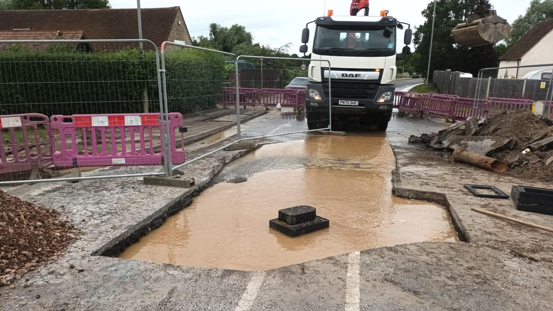 A road that has been dug up, showing a large amount of dirty brown water in it, with a lorry behind it. There are barriers, metal and plastic, and rows of rubble and mud piled up around it.