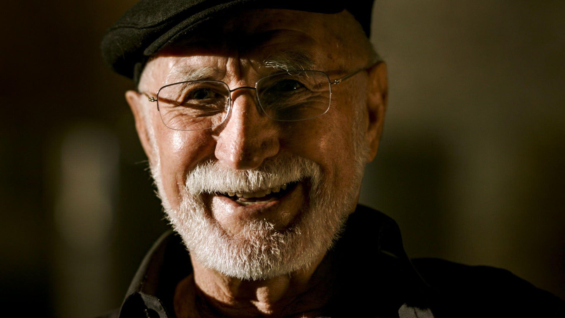 Roger Reeves who has glasses, a white beard and is wearing a black flat cap looking at the camera and smiling.