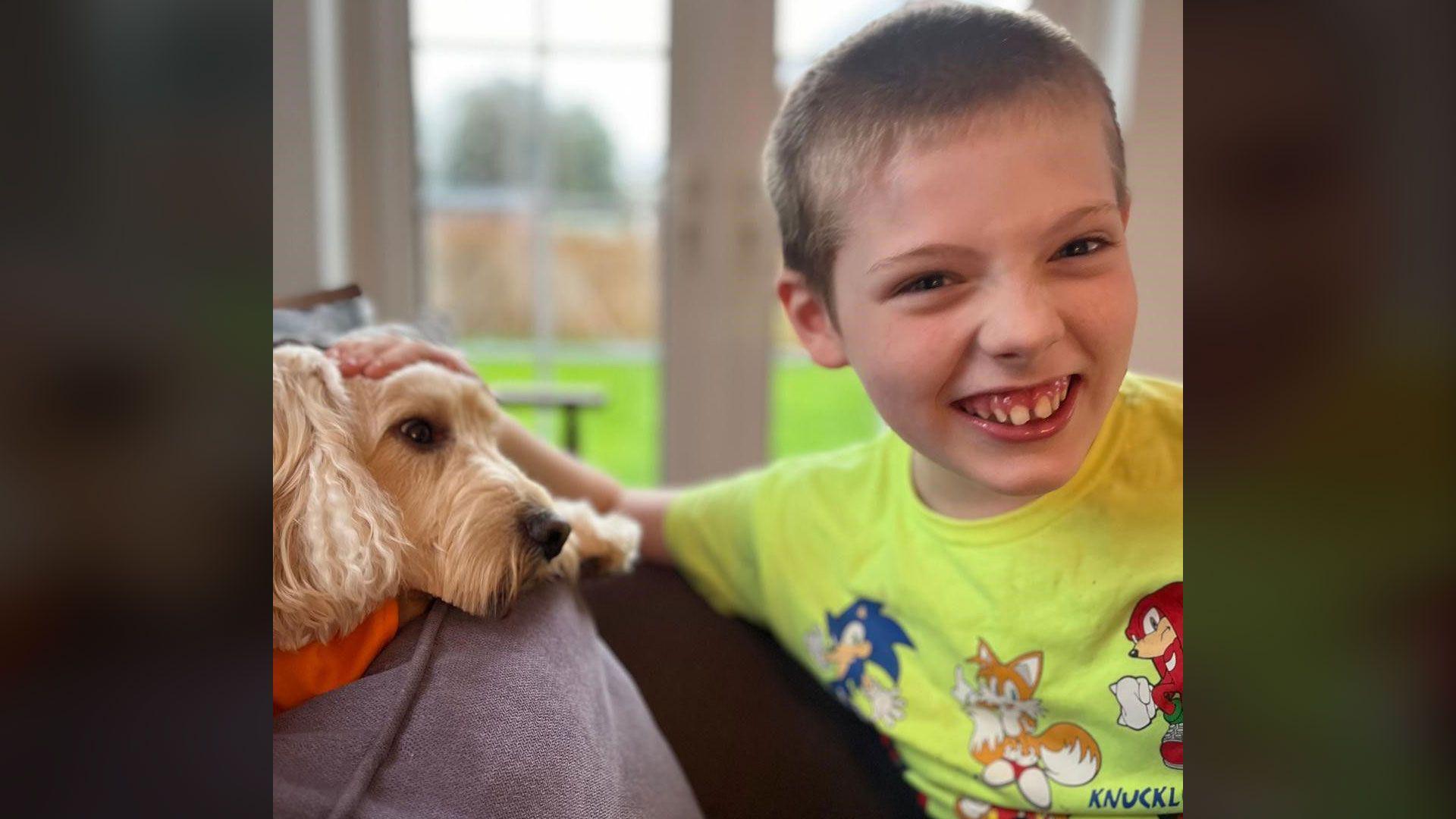 A young boy with blonde hair is laughing and looking into the camera. He is wearing a neon t-shirt with cartoon characters printed on the front. Beside him is a light brown dog lying on the back of a grey sofa. 