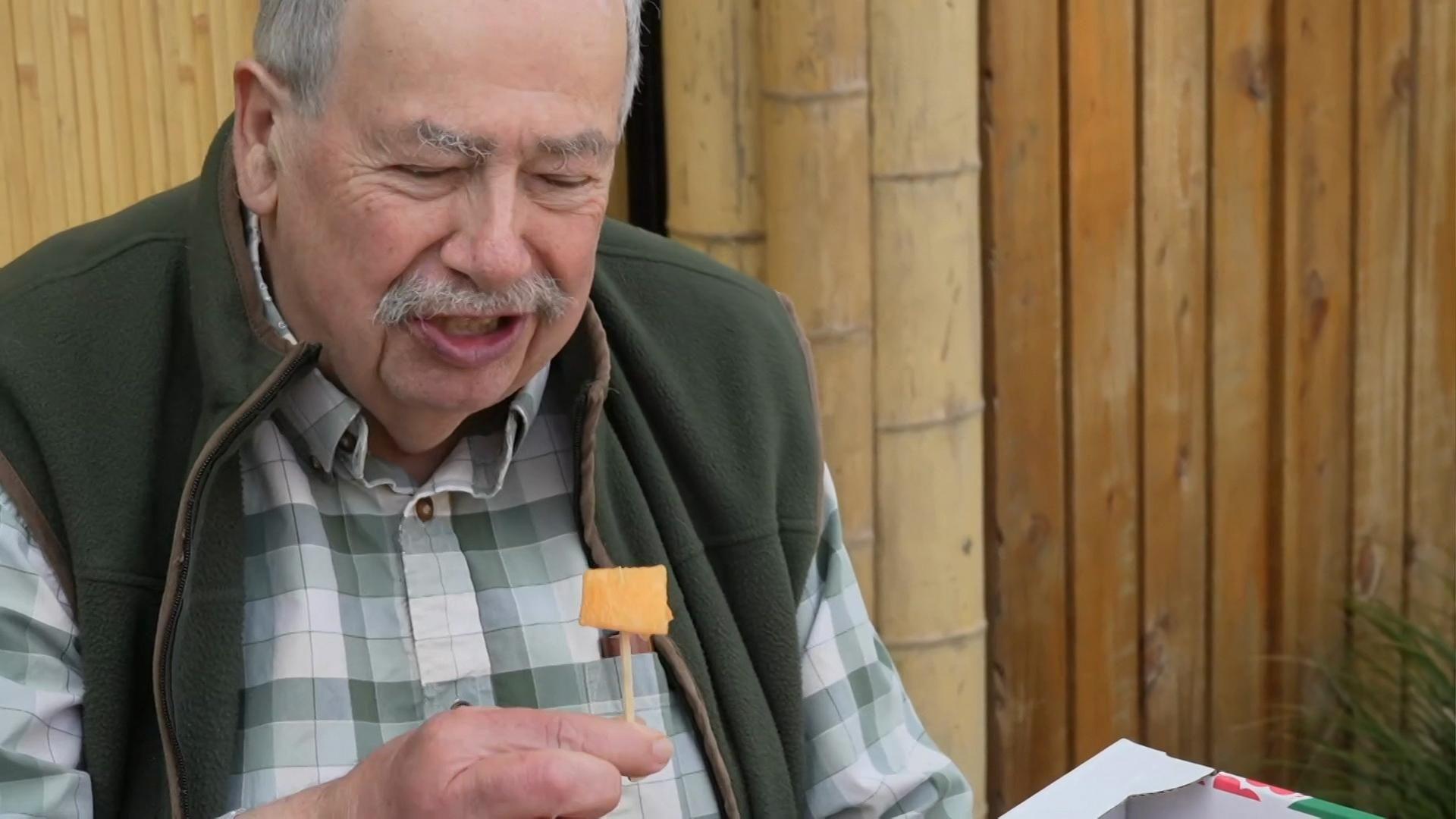 A man dressed in a shirt and overcoat holds a melon on a stick to taste test it