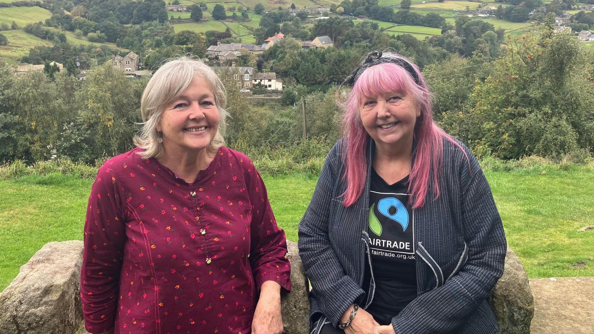Two women sit on a stone wall. The woman on the left is wearing a red dress and has shoulder-length grey hair. The woman on the right is wearing a black T shirt with a Fairtrade logo and a navy jackets. She has long pink hair.