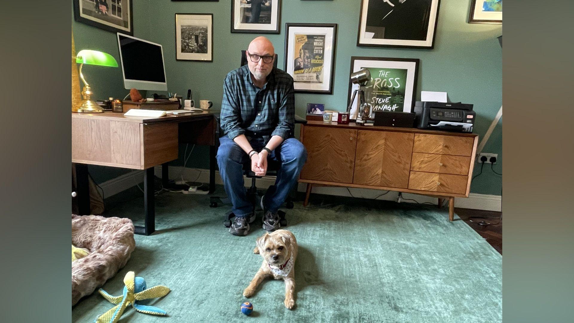 A man is sitting on a chair in an office, infront of him a small dog lies on the carpet with a ball