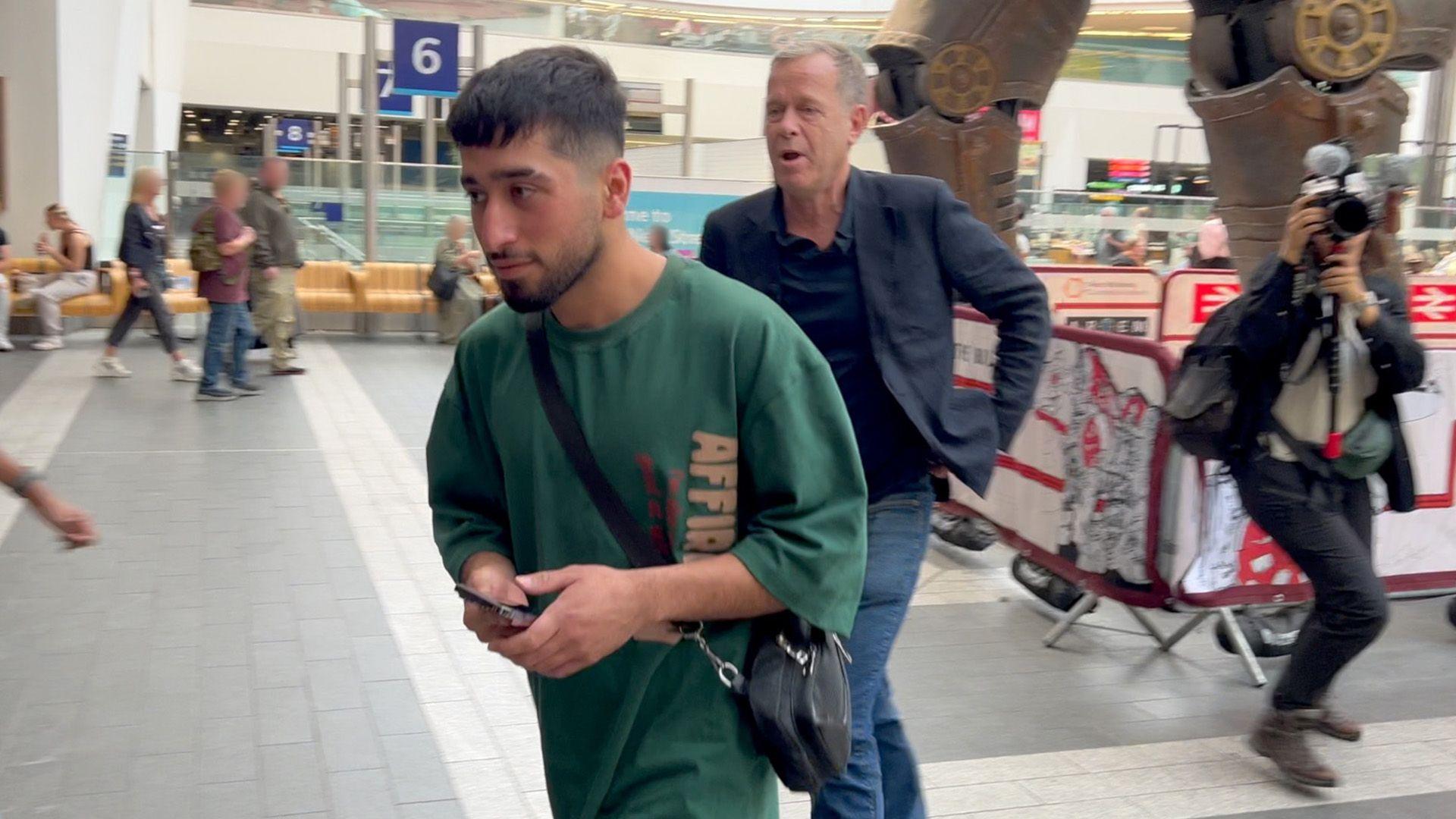 A young bearded man has turned away from journalist Andrew Harding and is heading to the Birmingham New Street station exit. The legs of the mechanical bull scuplture can be seen in the background and also in shot is a BBC cameraperson. 