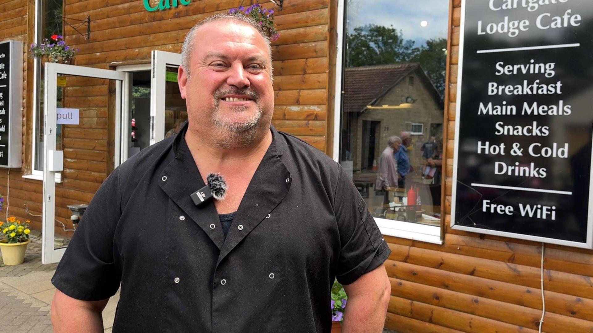 Adam Thurgood wearing a black chef shirt and a microphone on his lapel. He is standing in front of the wooden clad Cartgate lodge, which has a sign advertising breakfast, main meals, snacks, hot and cold drinks, and free Wi-Fi.