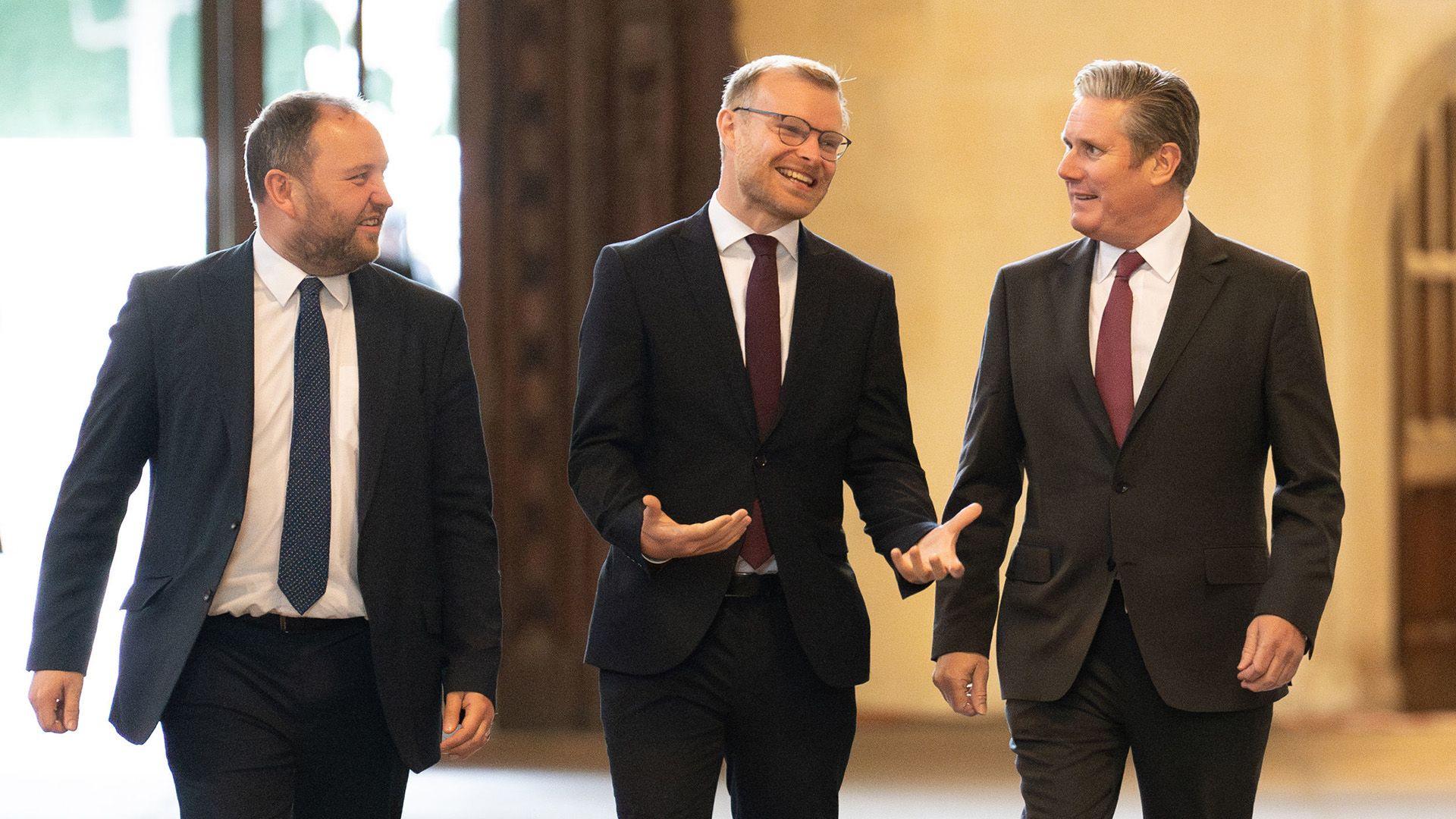 Murray, shanks and starmer walk along a corridor in a row wearing dark suits and ties.