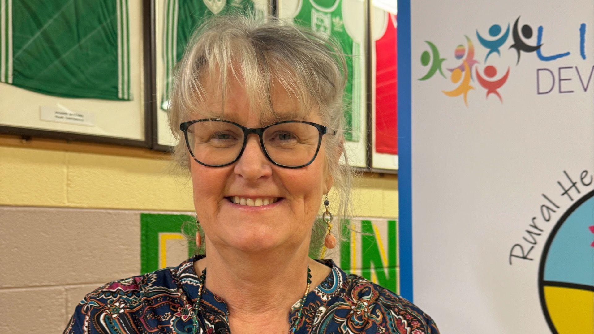 Marie McLoughlin, she has grey hair and is wearing black-rimmed glasses and a patterned top. She is standing in front of a row of framed boxing jerseys which are green and red.