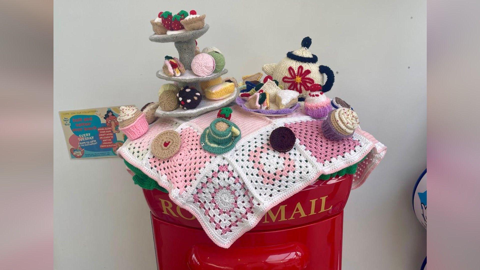 A knitted cake topper with knitted cakes, cake stands, and a white and red tea pot placed on a white and pink knitted napkin, all placed over the red post box