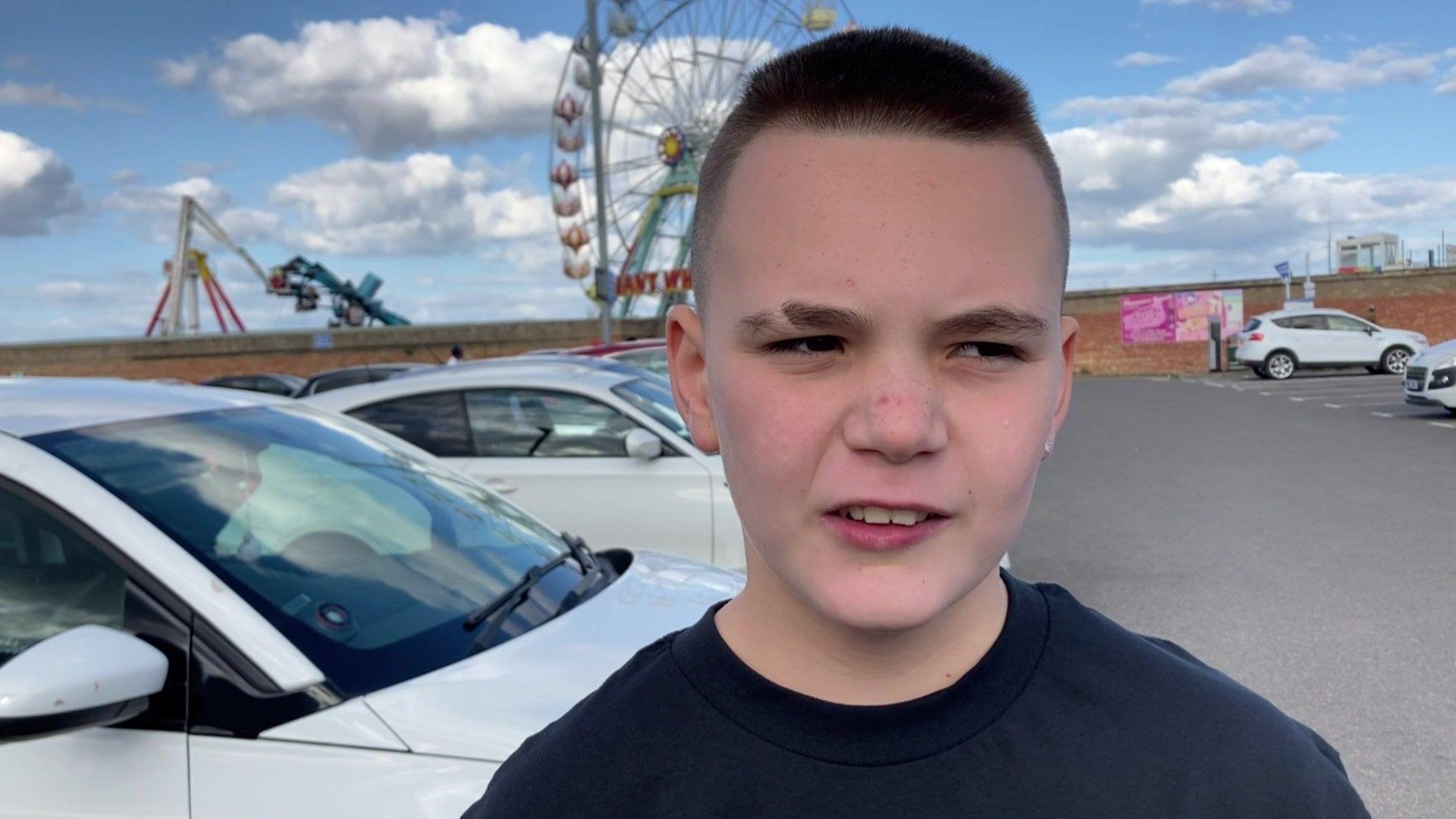 Young boy wearing a blue top with short dark hair stands in the car park of the fairground with cars in the background and the malfunctioned ride behind him