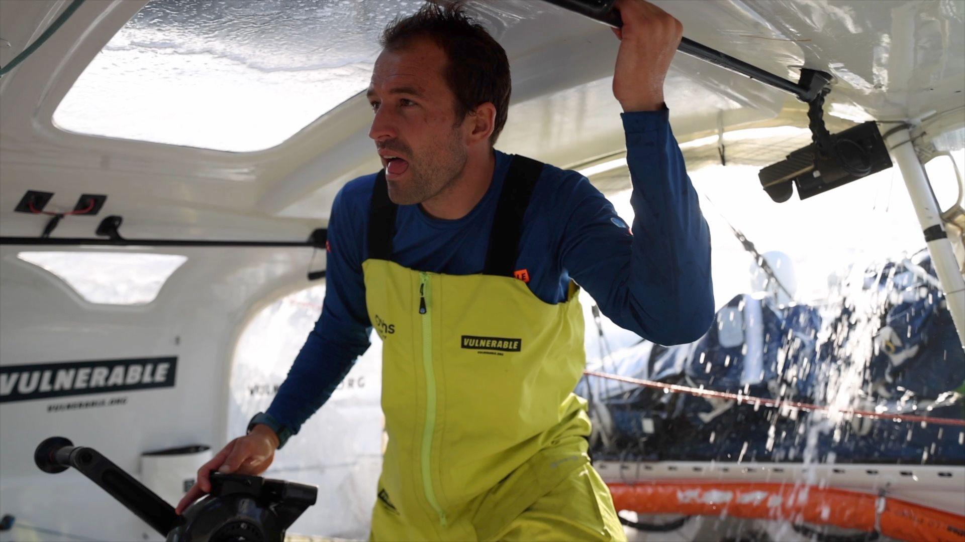 A man in a yellow sailing trousers and a blue top in a cockpit of a yacht with water pouring off the top of the cabin behind.