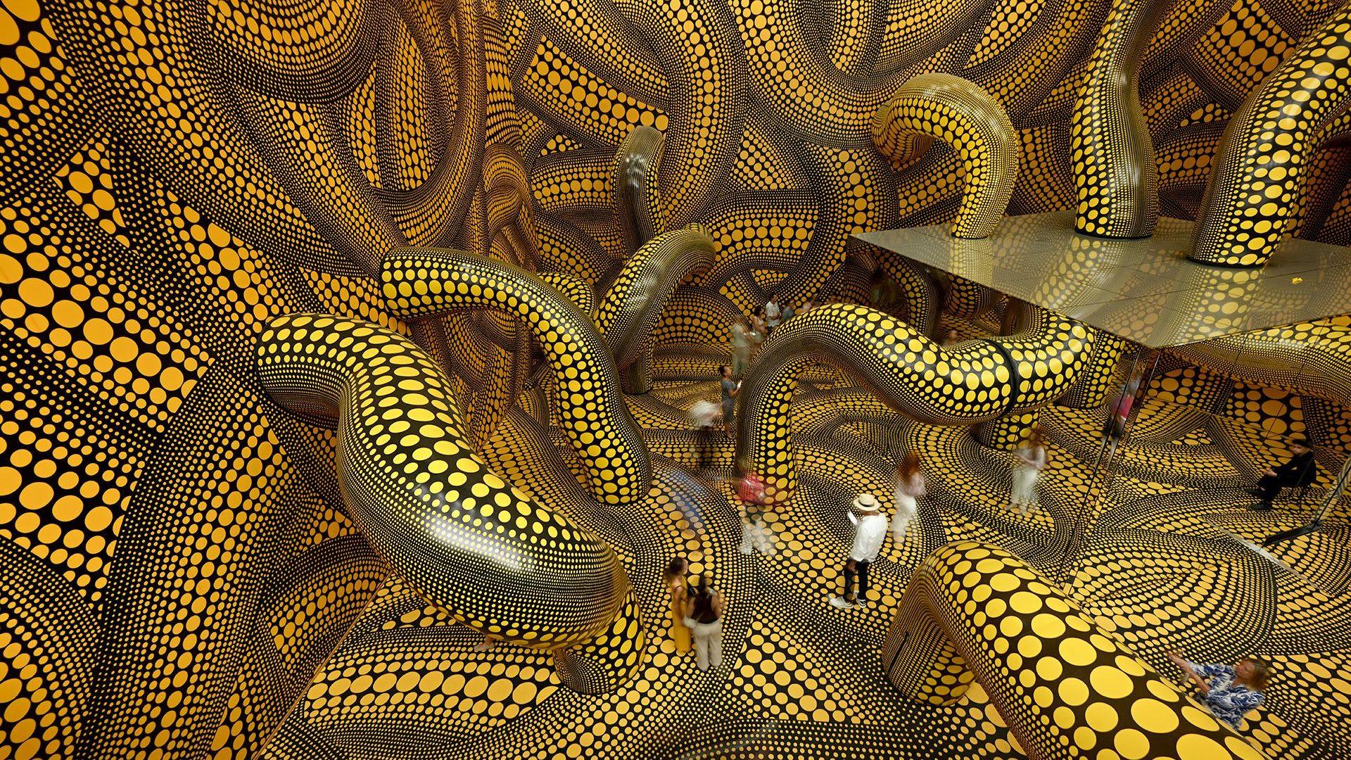 A series of intertwining tentacles in black and yellow polka dot run through a large room with mirrors in the centre, with visitors walking through it, in Basel in 2025.