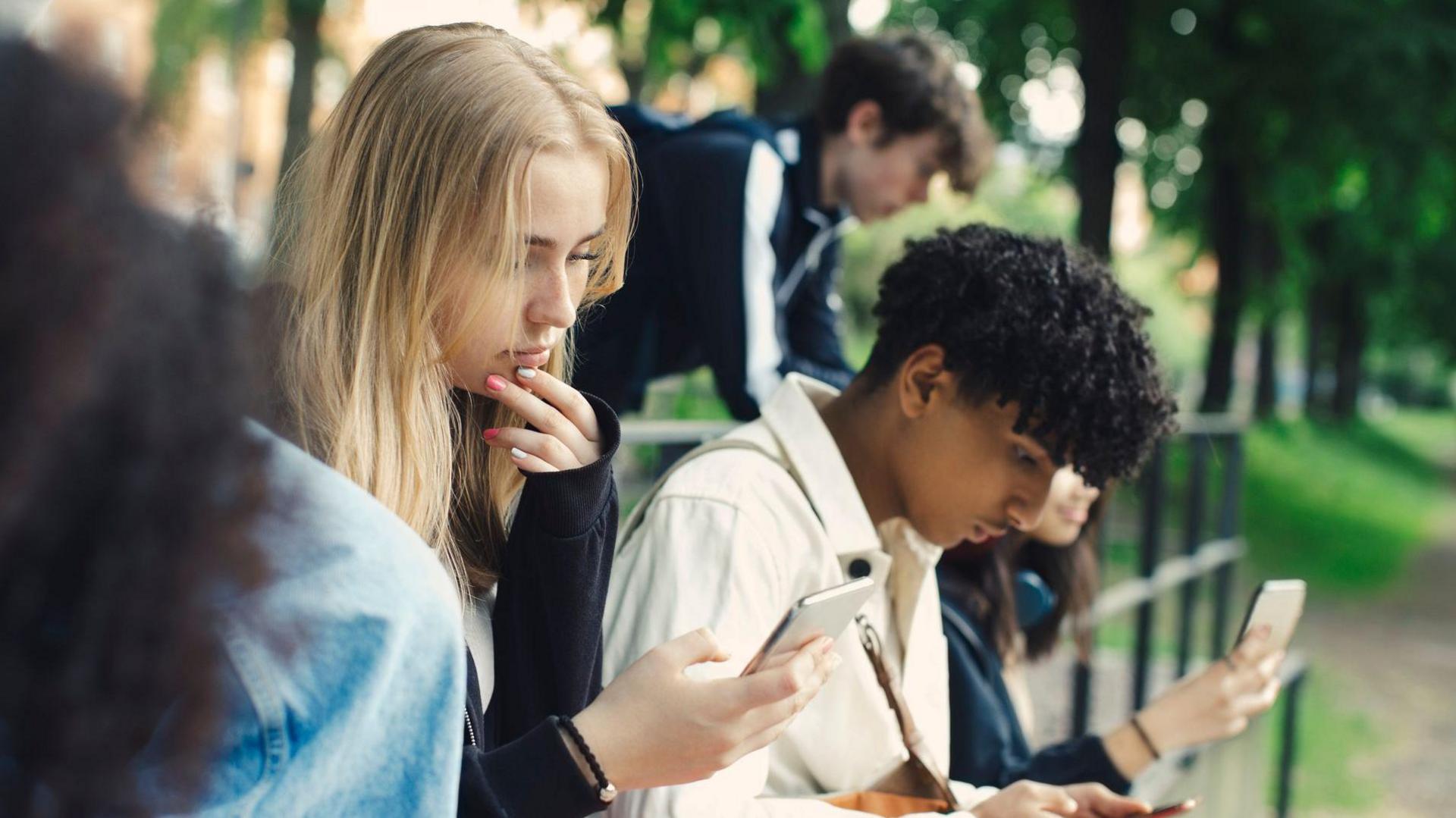 A group of teens look down at their smartphones in a park.