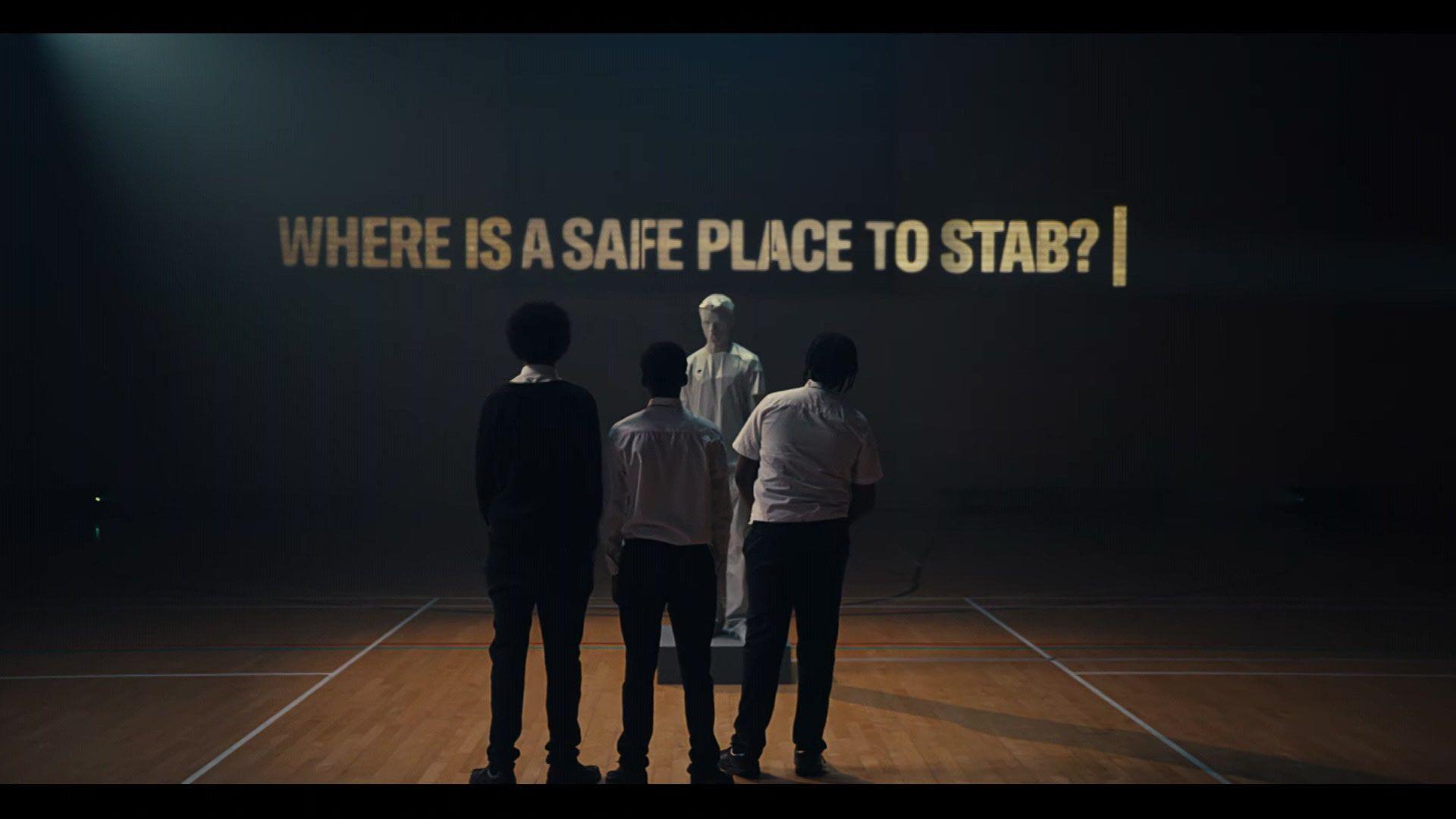 In a still from the campaign film, three teenage boys stand with their backs to us in a sports hall, looking at a male mannequin, and a film screen which bears the words 'Where is a safe place to stab?' 