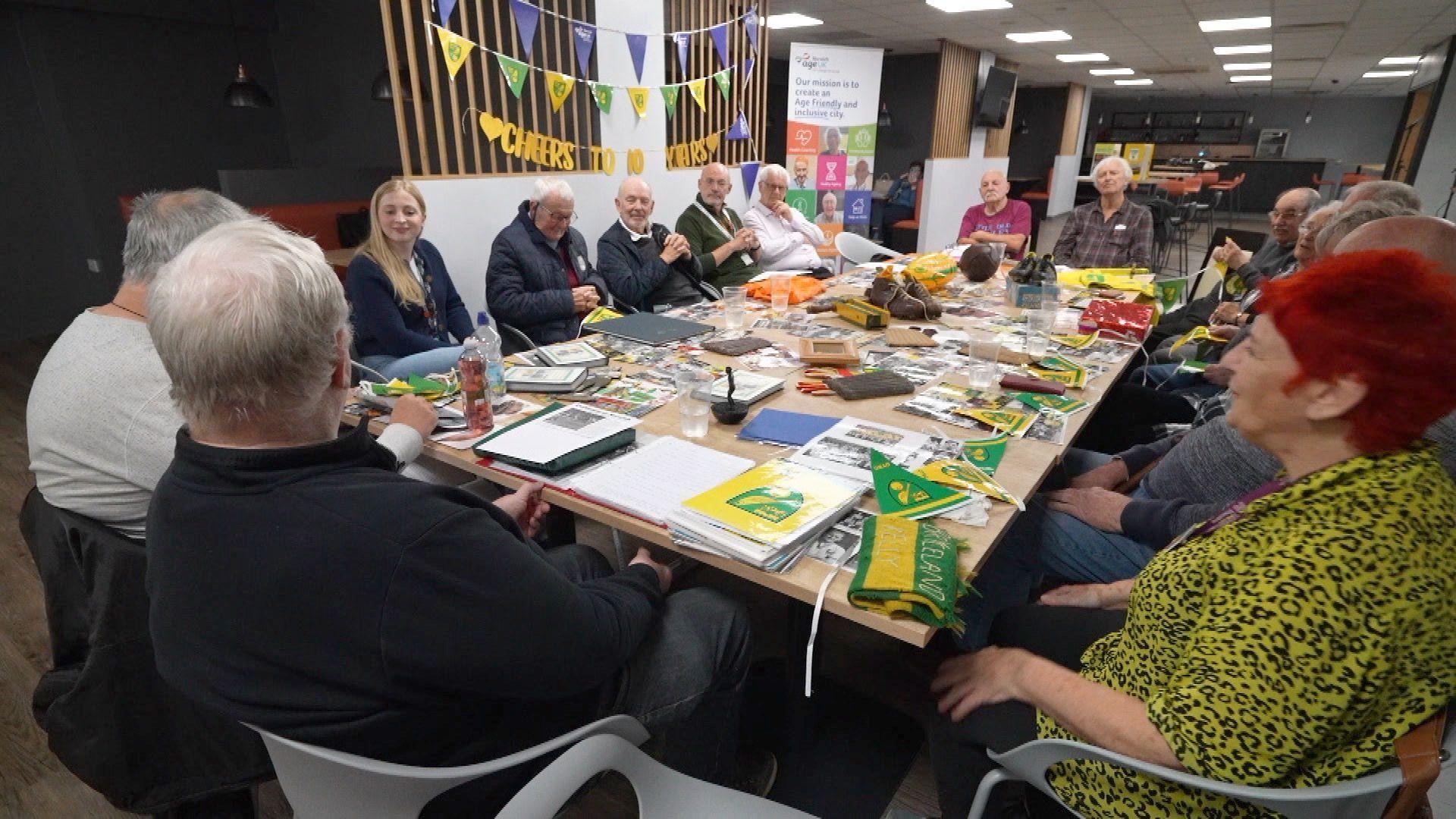 A group of people sitting around a table covered in Norwich City memorabilia. There are flags, old football boots, and pictures of former players. A woman in a yellow top sits at the front, and a man in a black shirt is also seated. They are in a function room.