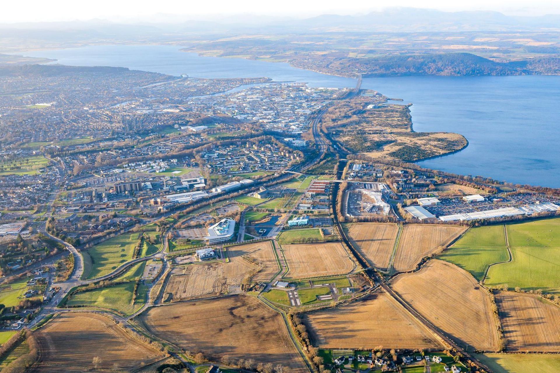 An aerial view of the east side of Inverness, including Inverness Campus, which is set in an area of brown-coloured fields. Also in shot are the Beauly and Cromarty firths and the Black Isle.