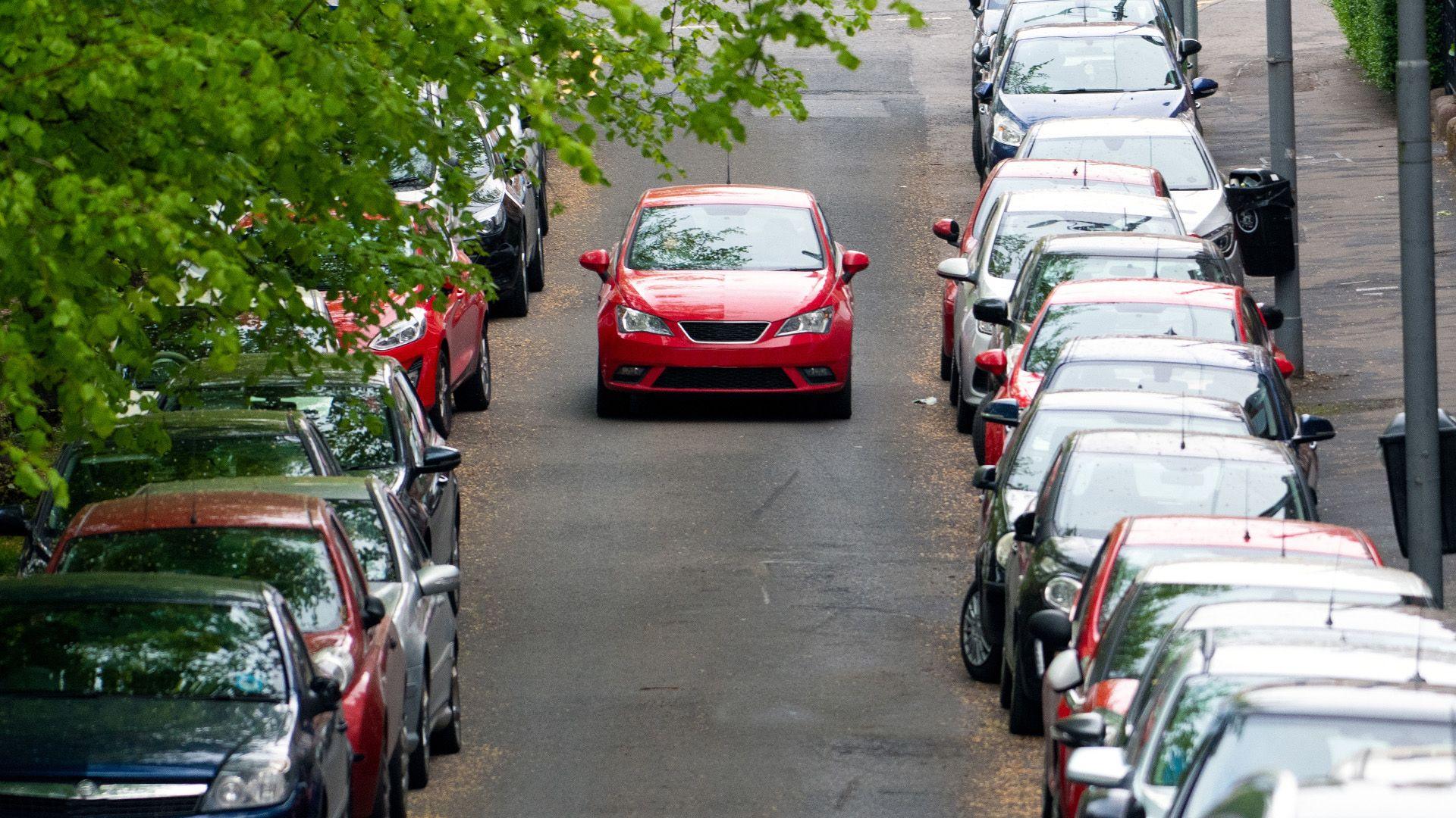 A red car drives down a road where lines of cars are parked on either side