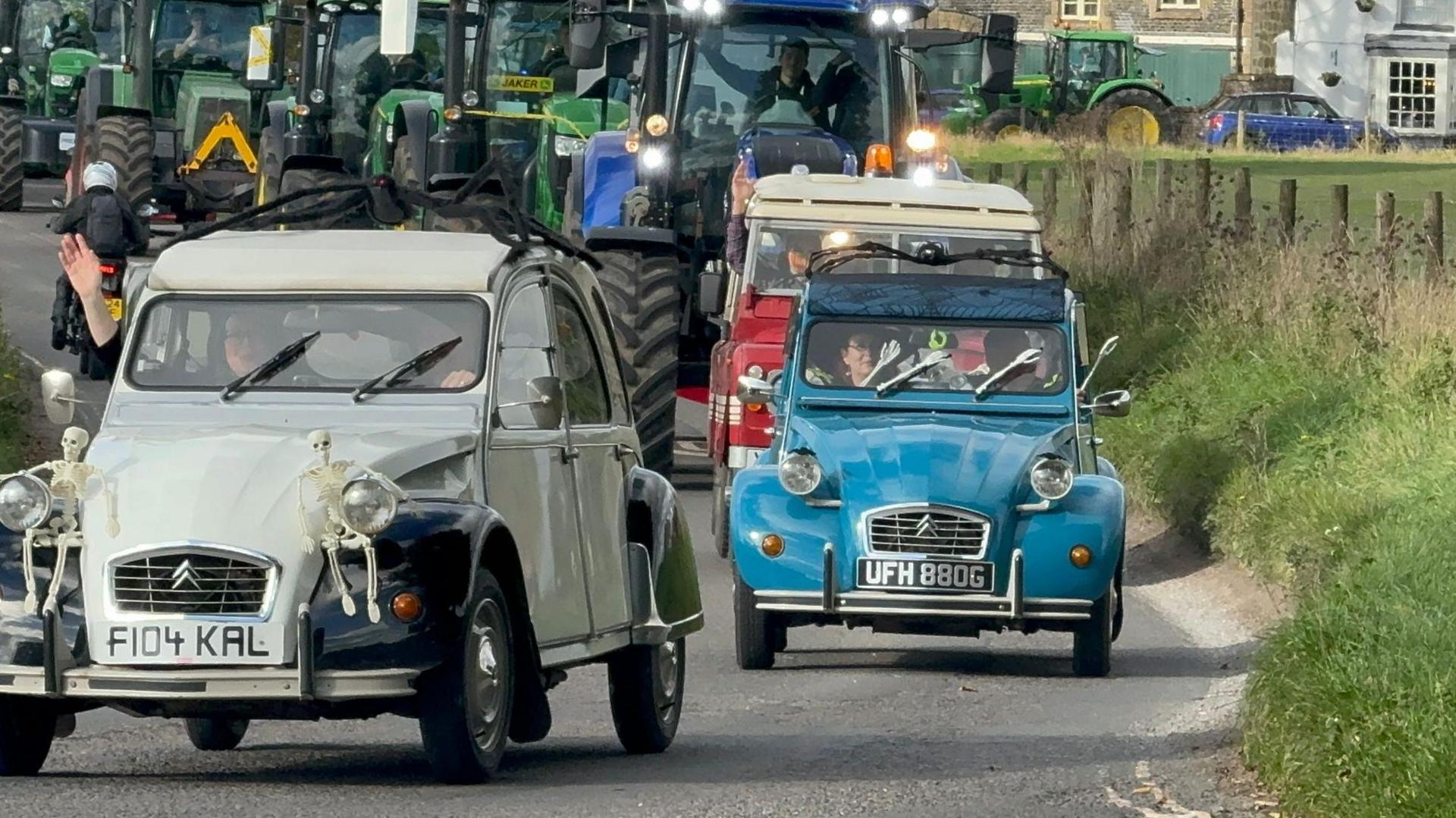 A cream and blue Citroen 2CV, followed by a red military vehicle, head up a convoy of tractors driving down a country road.  