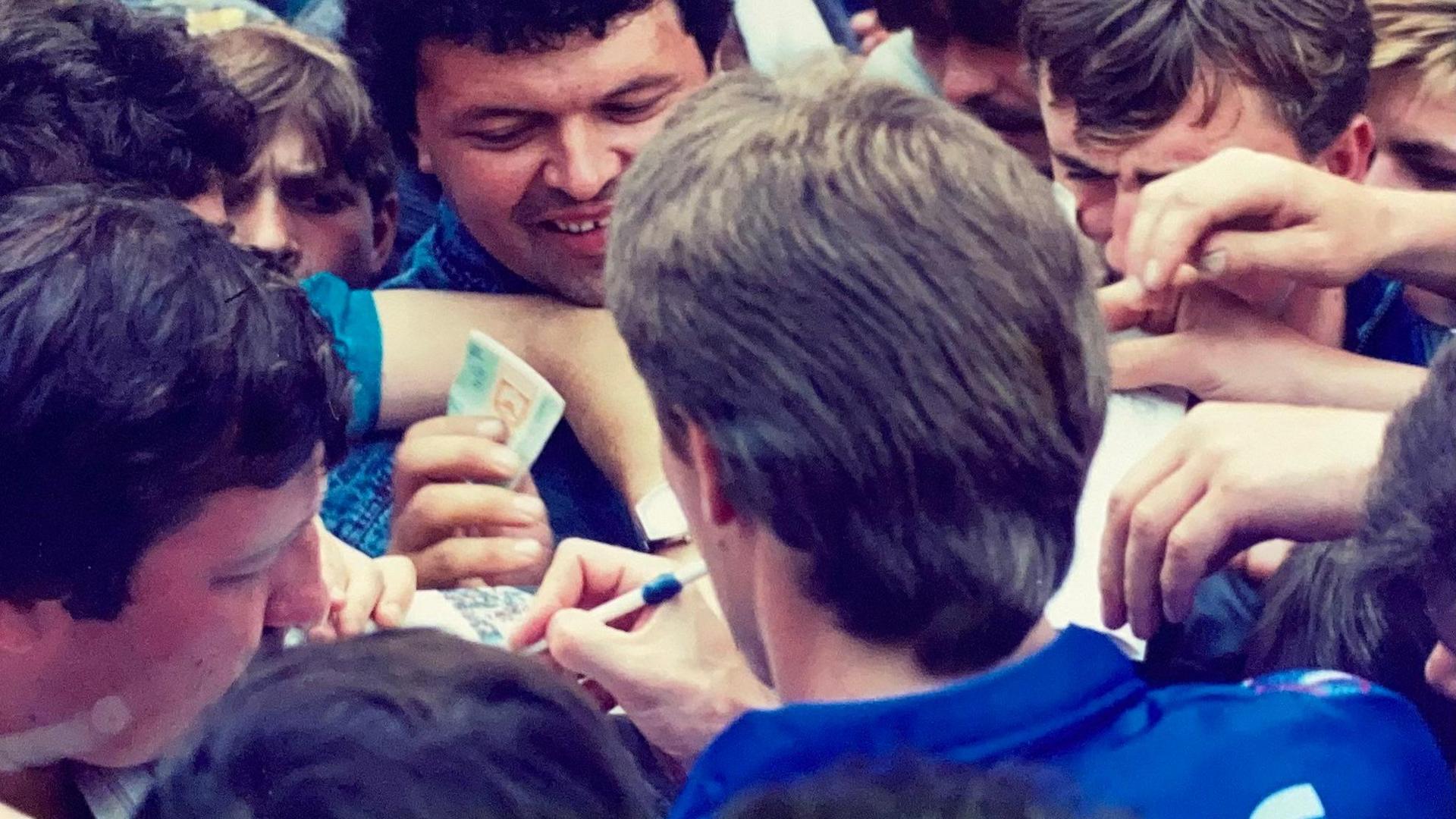 Sergei Baltacha is pictured from behind signing autographs for a busy crowd os people. He has thick hair swept back and is wearing a blue top.