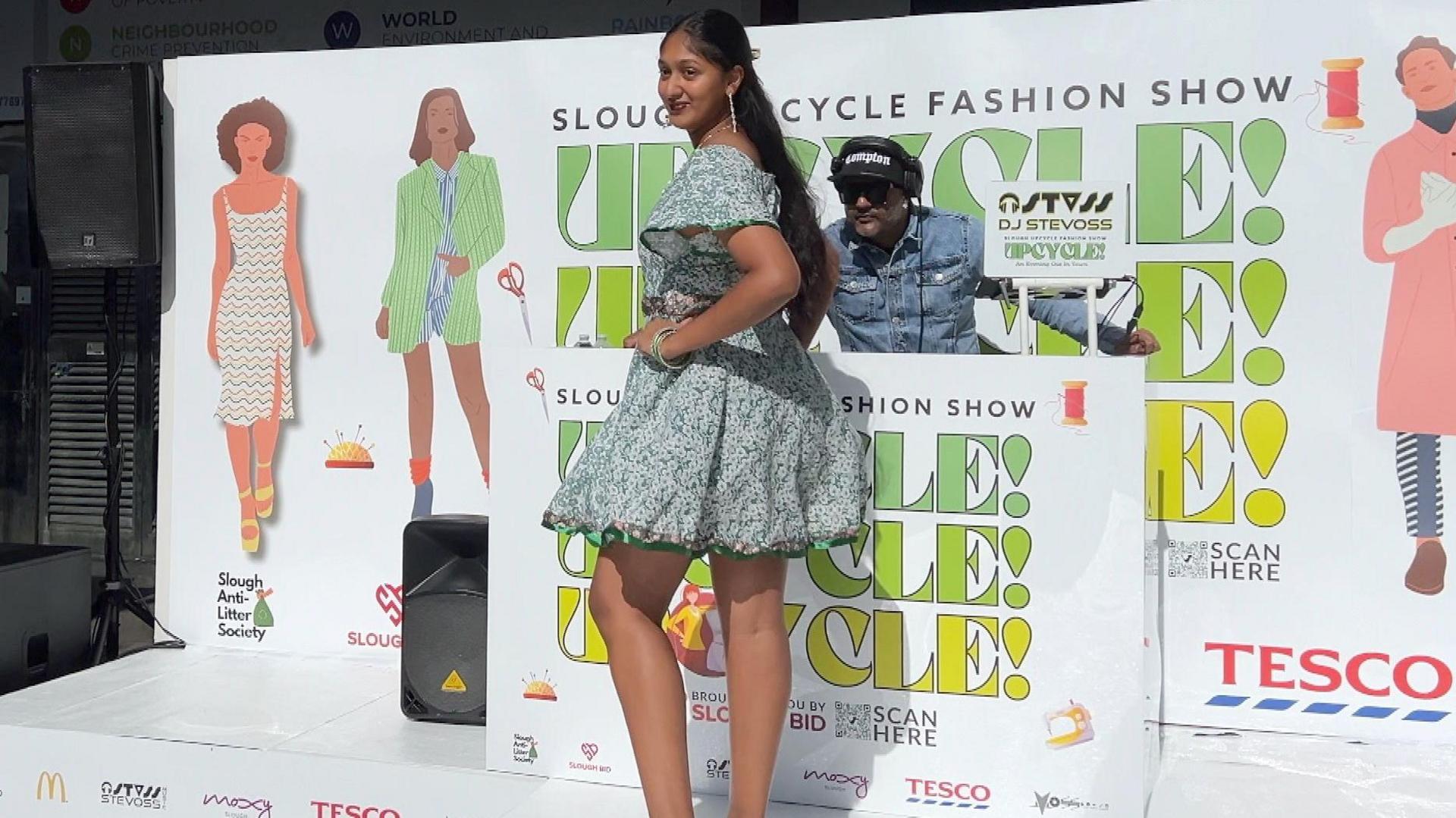 A woman with long dark hair poses on the catwalk wearing a short light green patterned dress.