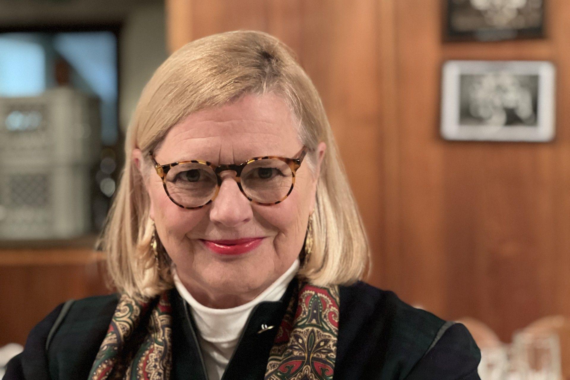 An older woman with shoulder length blonde hair is wearing glasses, a white polo neck and black jacket and a patterned scarf. She is in a wood-panelled room with three framed images on the wall.