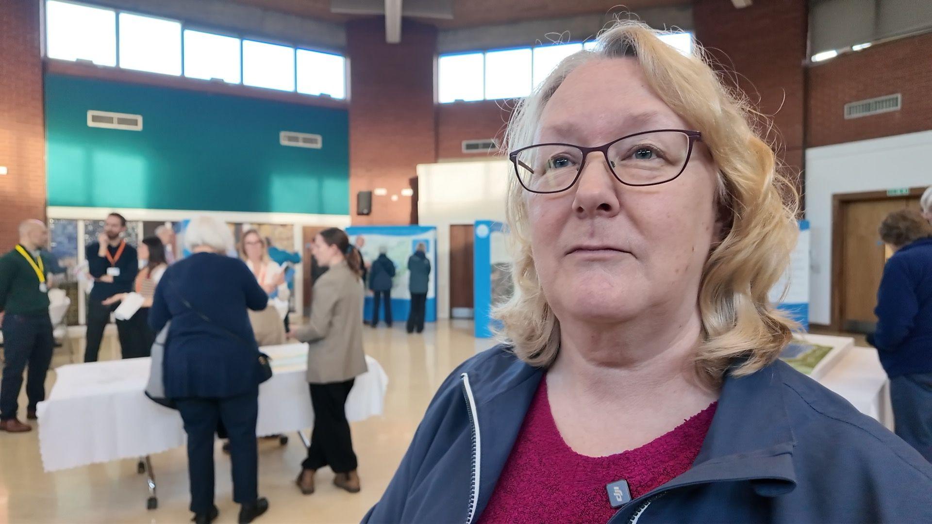 A woman with mid length wavy hair and glasses in a blue jacket stands in the hall containing the reservoir exhibition