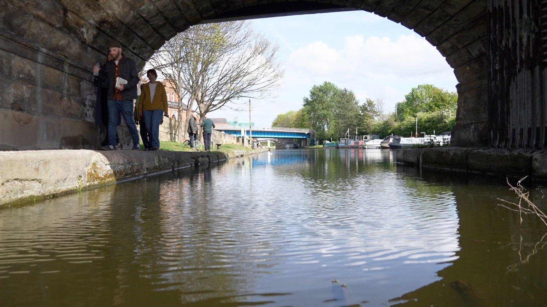 The Bridgewater Canal shown through a tunnel, with a small group of people walking alongside the water. Through the tunnel, a bridge and a small number of boats are seen in the distance.