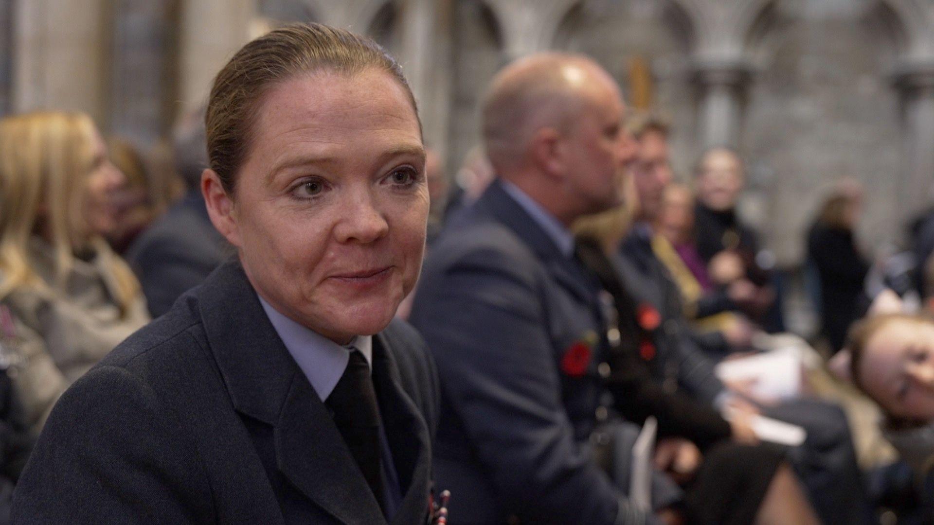 Nick Startup wears an army uniform with a blue shirt and tie. Her hair is tied back. Other people in uniform are sitting int he pews at the side of her.