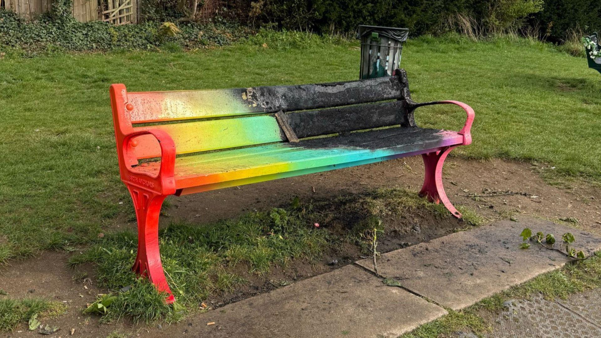 A park bench painted in various colours which has been set on fire as half of it is blackened.