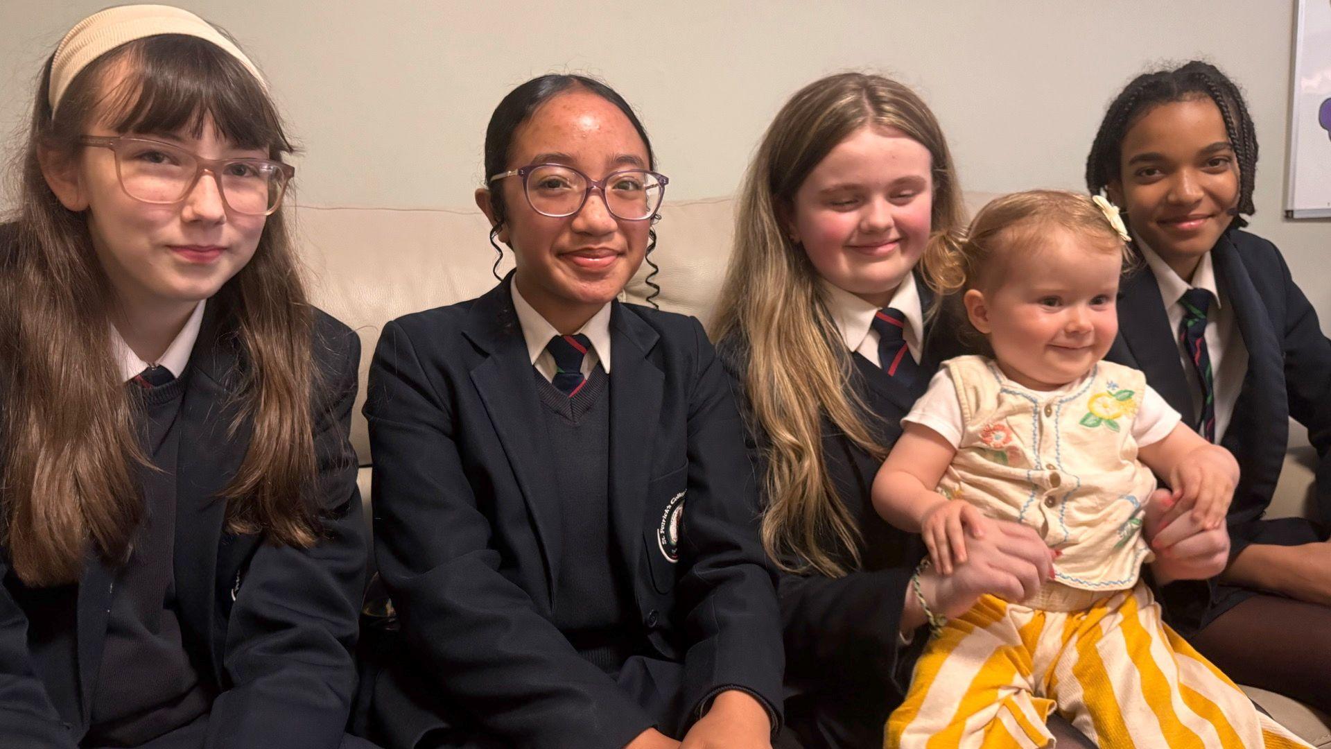 Four teenage schoolgirls and a young girl, aged about two - the schoolgirls are wearing dark school uniforms, with navy blazers and navy ties with a red stripe. The young girls has a yellow striped bottoms and a yellow vest. She has short blond hair set into short pigtails. 