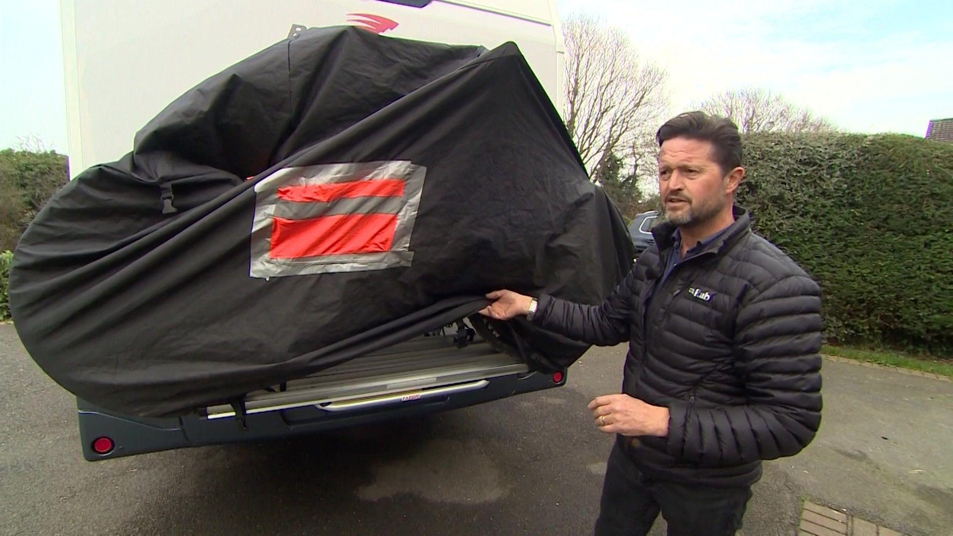 Adrian Fenton lifting the bike rack's cover up. It is on the back of the motorhome, which is parked on a driveway. Mr Fenton is wearing a black jacket and is talking.