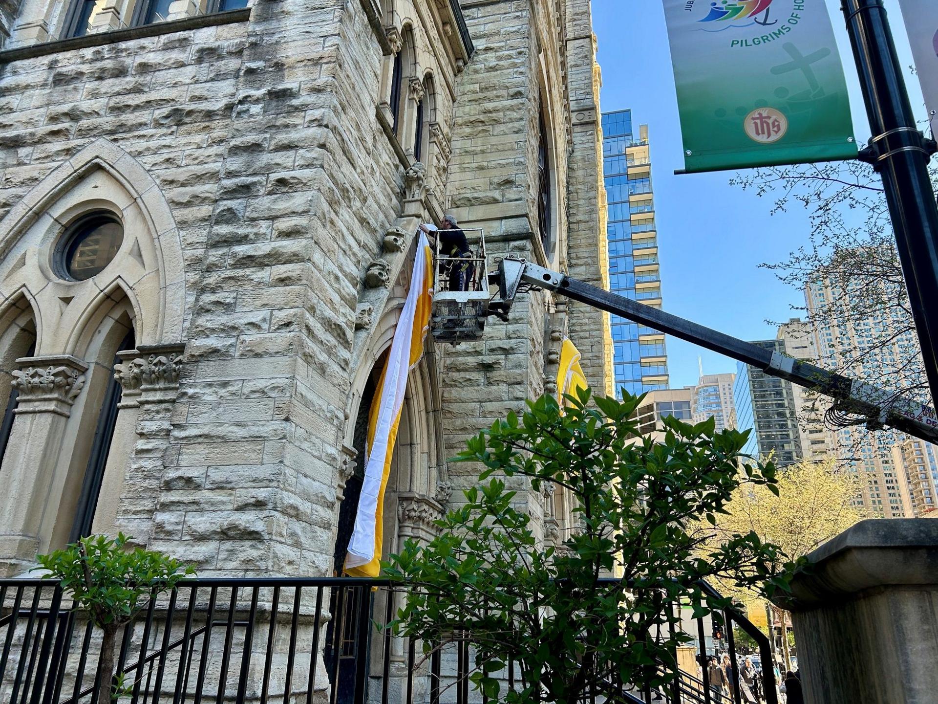 A stone building with more modern skyscrapers in the background, a man on a crane affixes a white and yellow banner to the outside of the church.