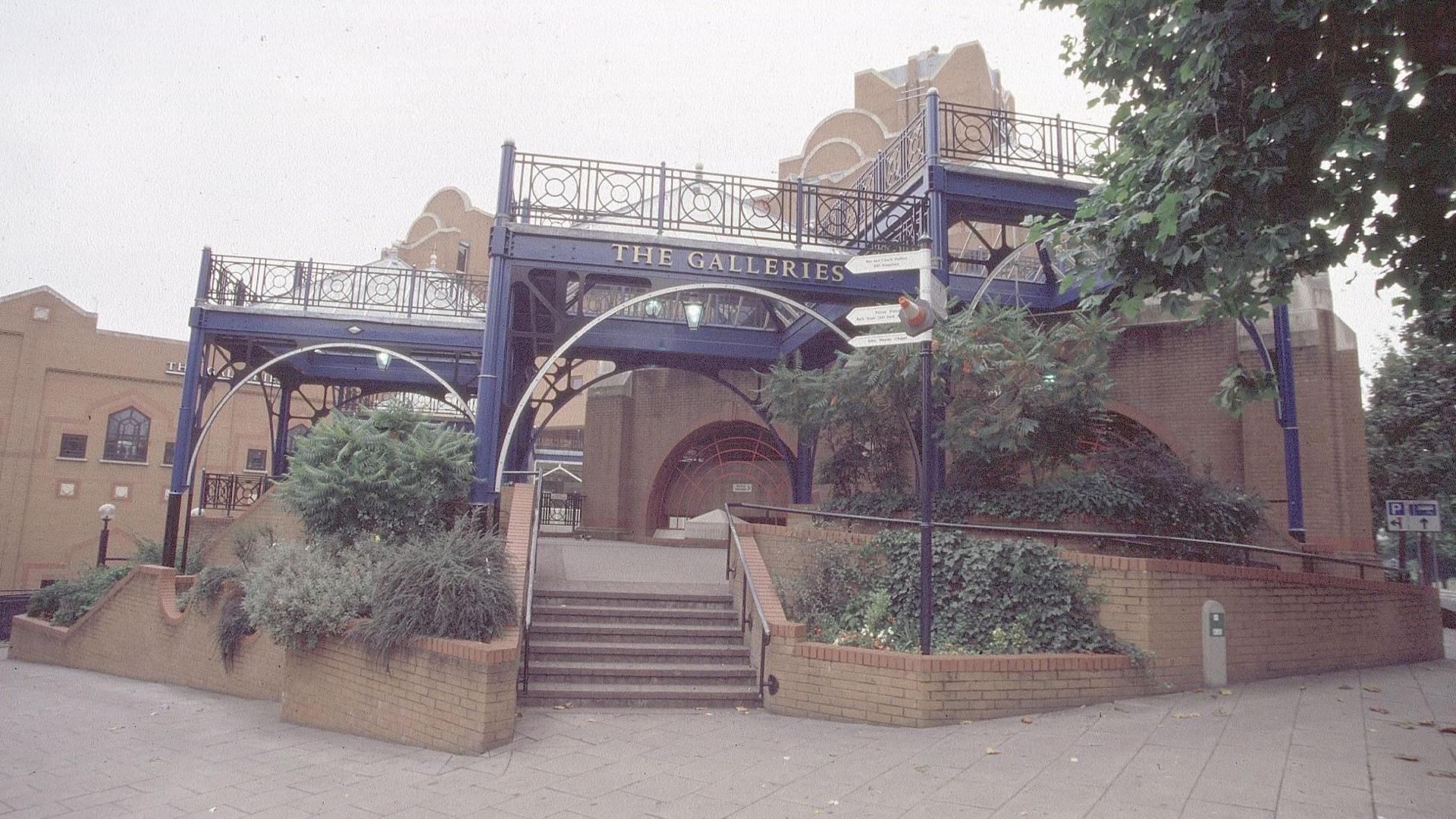 An image of one of the entrances to the Galleries as it currently looks. It has brick walls and steps running through blue railings, along with shrubbery. 'The Galleries' is seen on a blue arch over the entrance