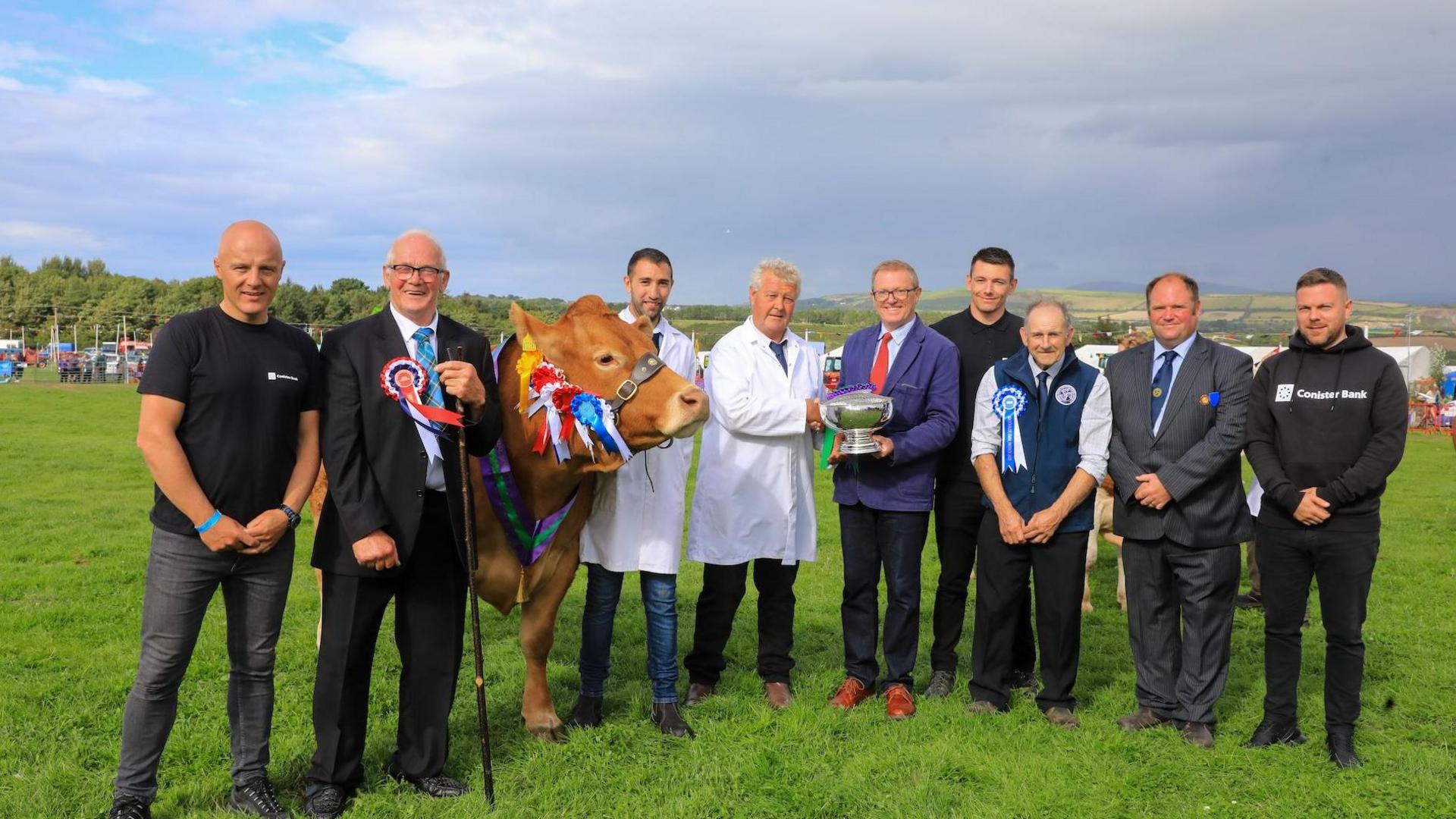 Nine men smiling in a field, two wear white coats, and a cow with a number of rosettes stands among them.