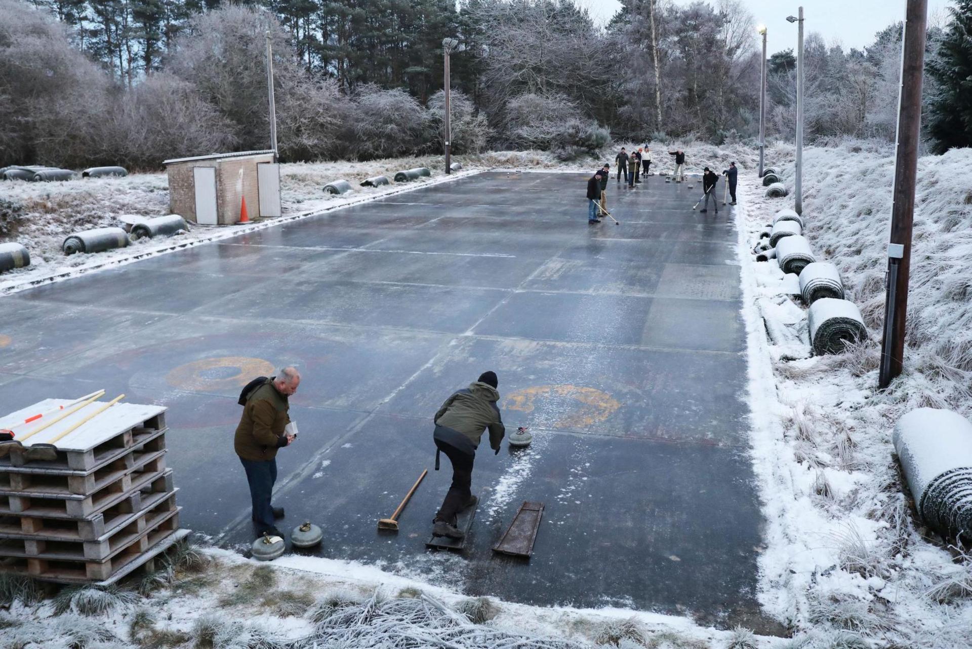 Curlers in action at the rink in Muir of Ord. There is snow lying all around in the rink. There are large rolls of coverings for the rink.