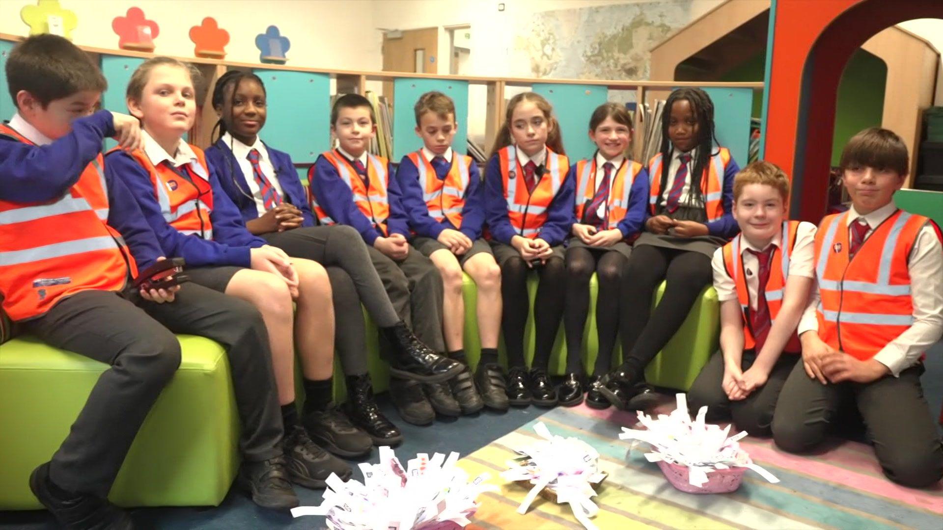 A group of school children in blue school uniform wearing orange hi-vis jackets. They are sat on a green curved sofa.