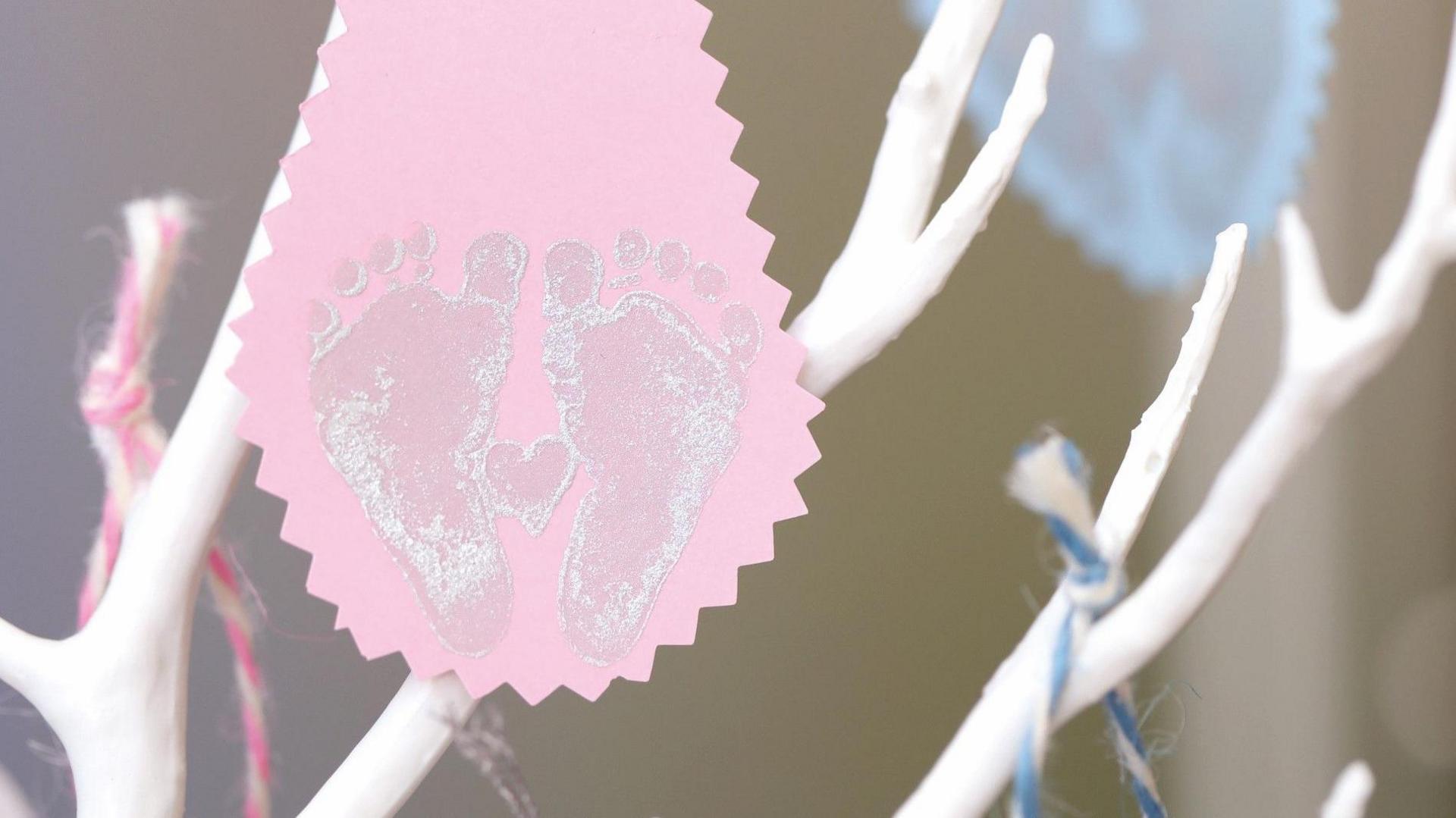 A pink tag with a painted stamp of baby's feet on it, places on a white wooden tree