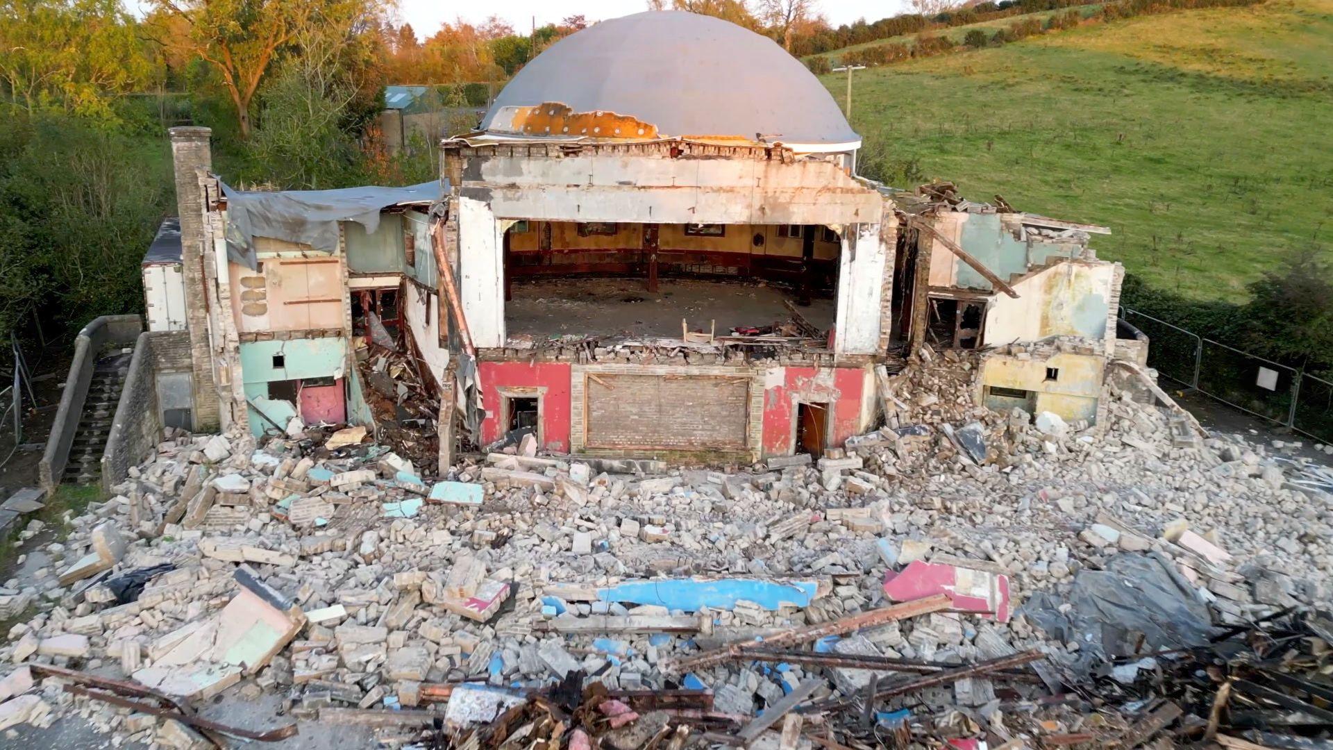 Edendork Hall with a lot of rubble on the floor and a frame of the building with the back wall still intact. The dome is also still up which is grey and has small specks of gold left on it. Behind the demolished building is a green field. The image has been taken during sunset so the grey building is reflected by a reddish sunlight. 