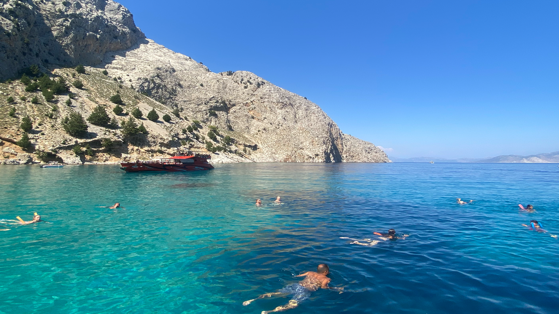 A turquoise sea below a light-coloured cliff face. In the sea, there are several people swimming, spread out across the image. In the background, there is a red, large boat. 