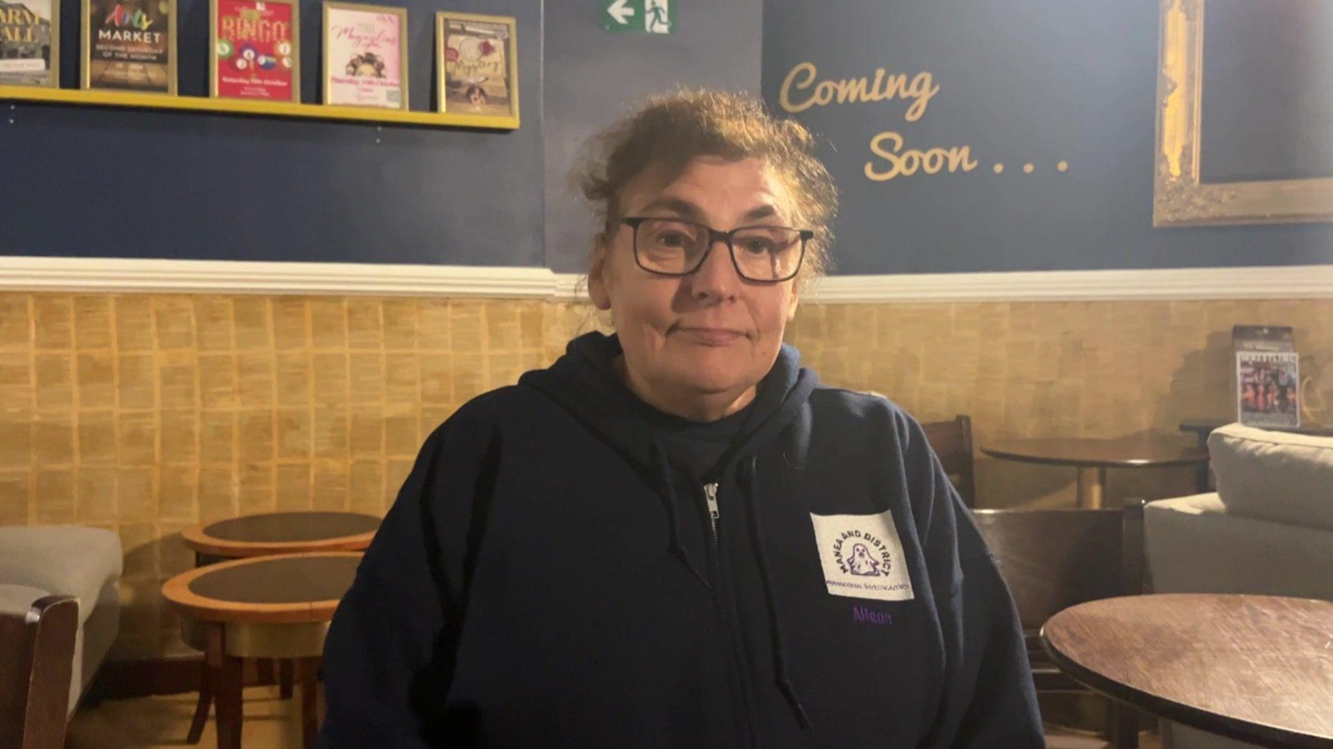 Alison Searle is wearing a navy zip-up hoodie with a logo on the top right. The logo is white with an outline of a ghost in the centre, above it reads "Manea and District Paranormal Investigators". She has her hair up, is wearing square-framed glasses and is smiling at the camera. She is sitting in what looks to be a café. There are several wooden tables and chairs behind her.