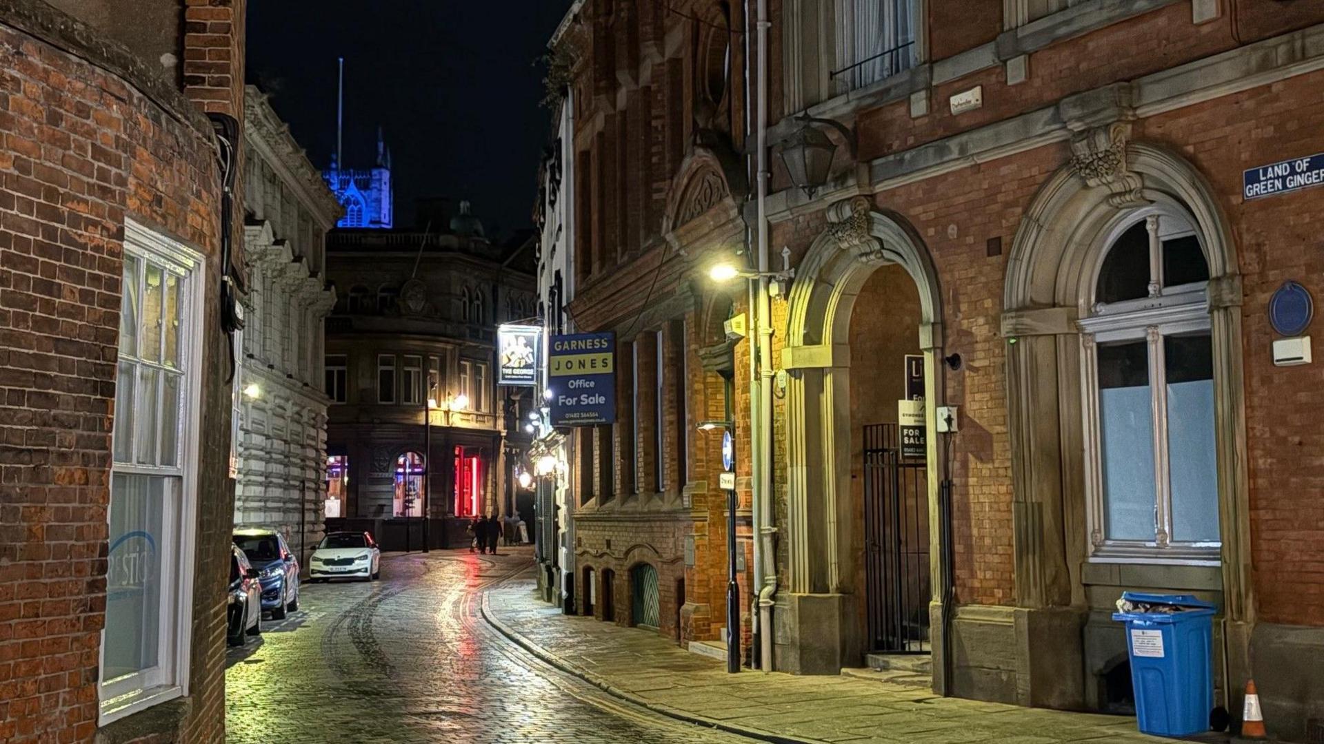 A photograph, taken at night, show a cobbled street curving to the right. On the inside bend is a brick-built Victorian-era building. The cobbles are glowing from the reflection of streetlamps.