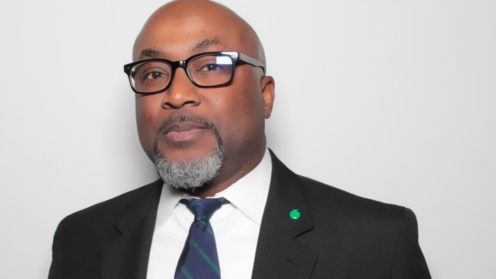 Aland Etienne, in black-rimmed glasses, is wearing a black suit jacket and white shirt with a dark blue and green patterned tie. He is standing against a plain white backdrop.