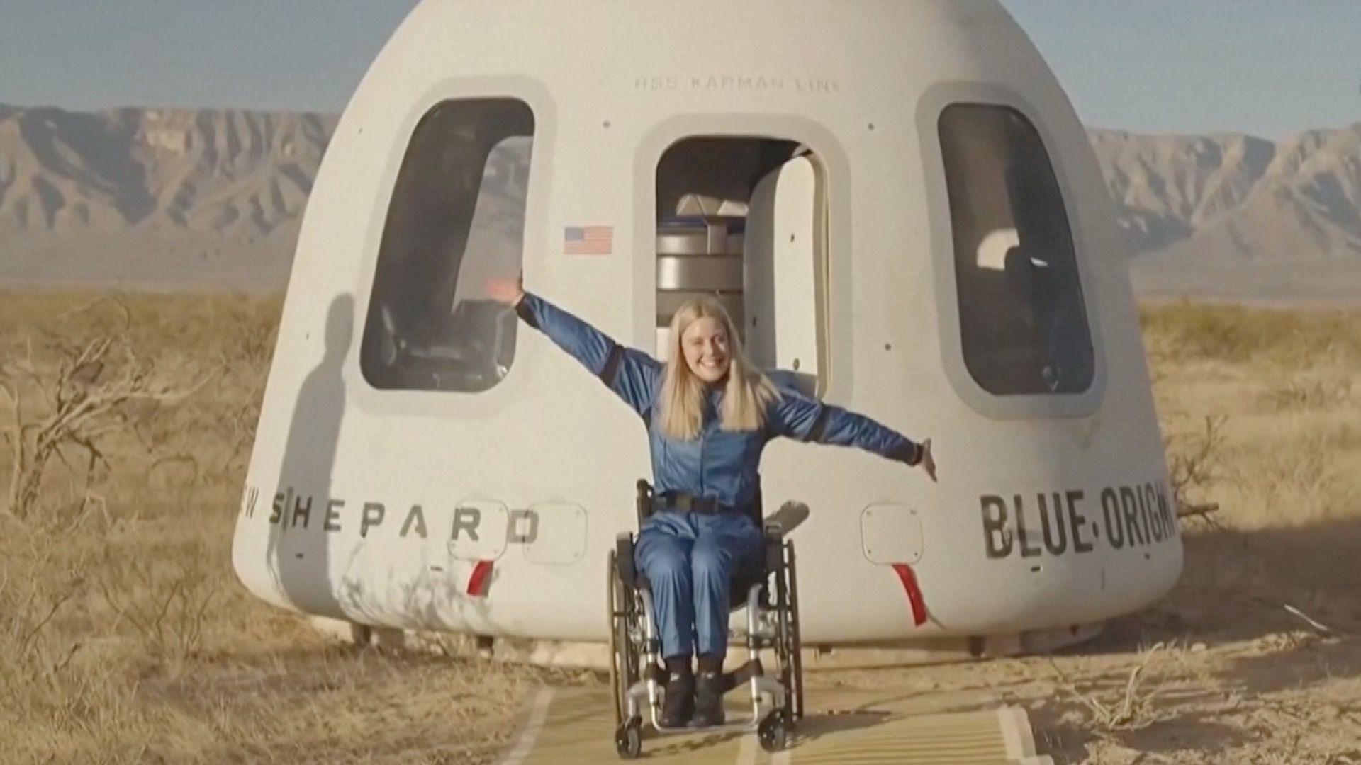 woman in blue boiler suit in wheelchair sits with arms out wide in front of Blue Origin capsule in a desert with mountains seen in the far distance