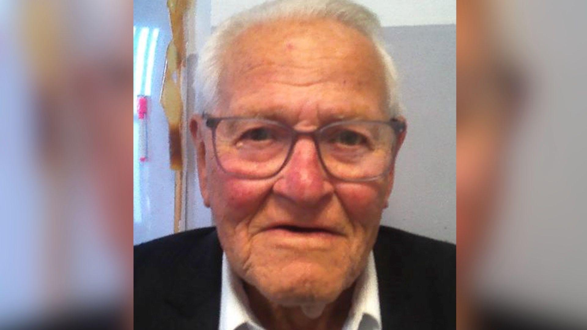 An elderly-looking man is staring at the camera with a neutral expression. He has thinning grey hair and glasses on. He's wearing a black jumper.