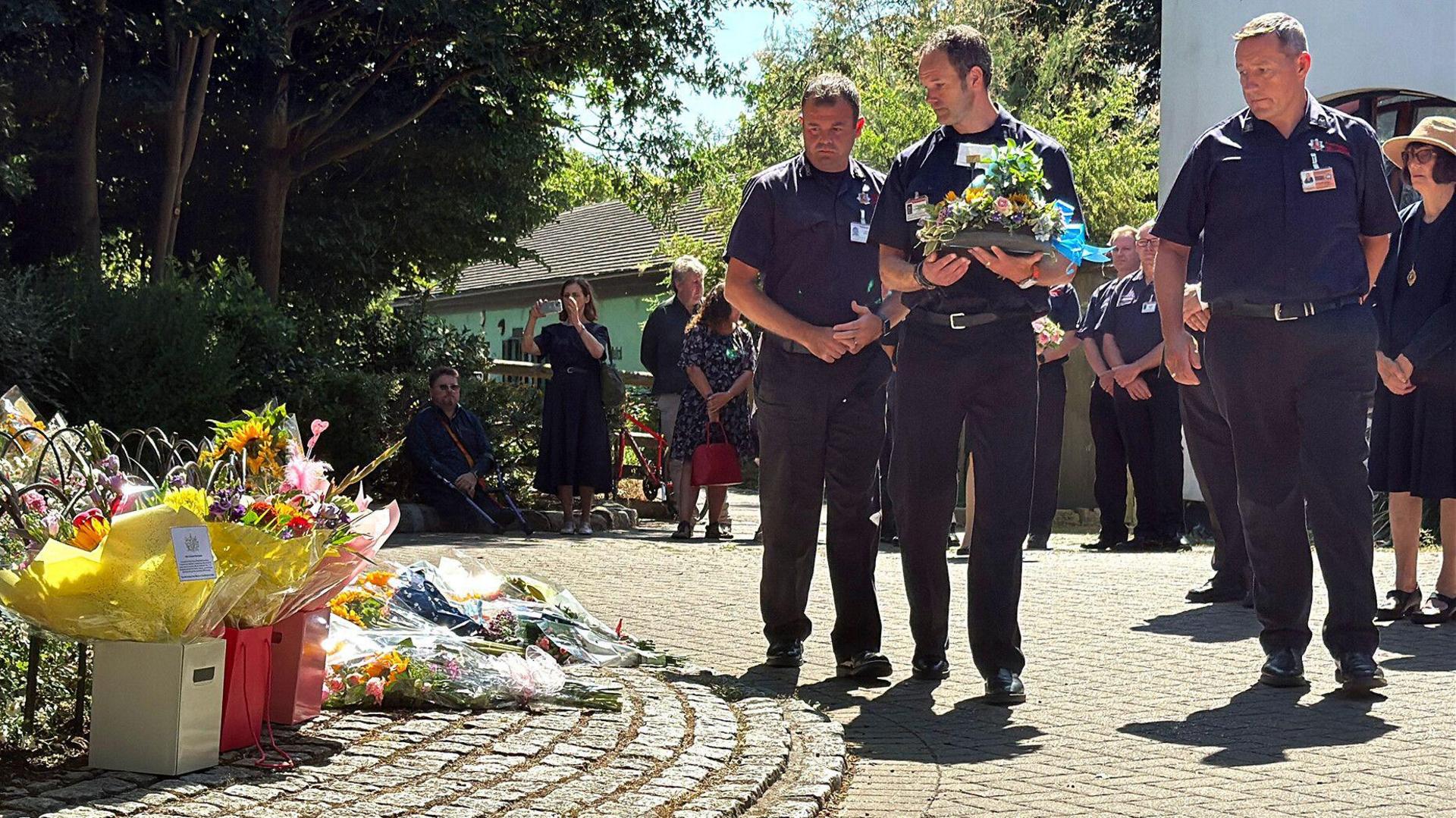Three male firefighters, one carrying a bouquet of flowers approach an area in the park where flowers and soft toys have been left in a circle in tribute to the deceased child Leonna Ruka who died after a falling tree branch collapsed on her. Several men and women can be seen standing nearby. It is a warm and sunny day.