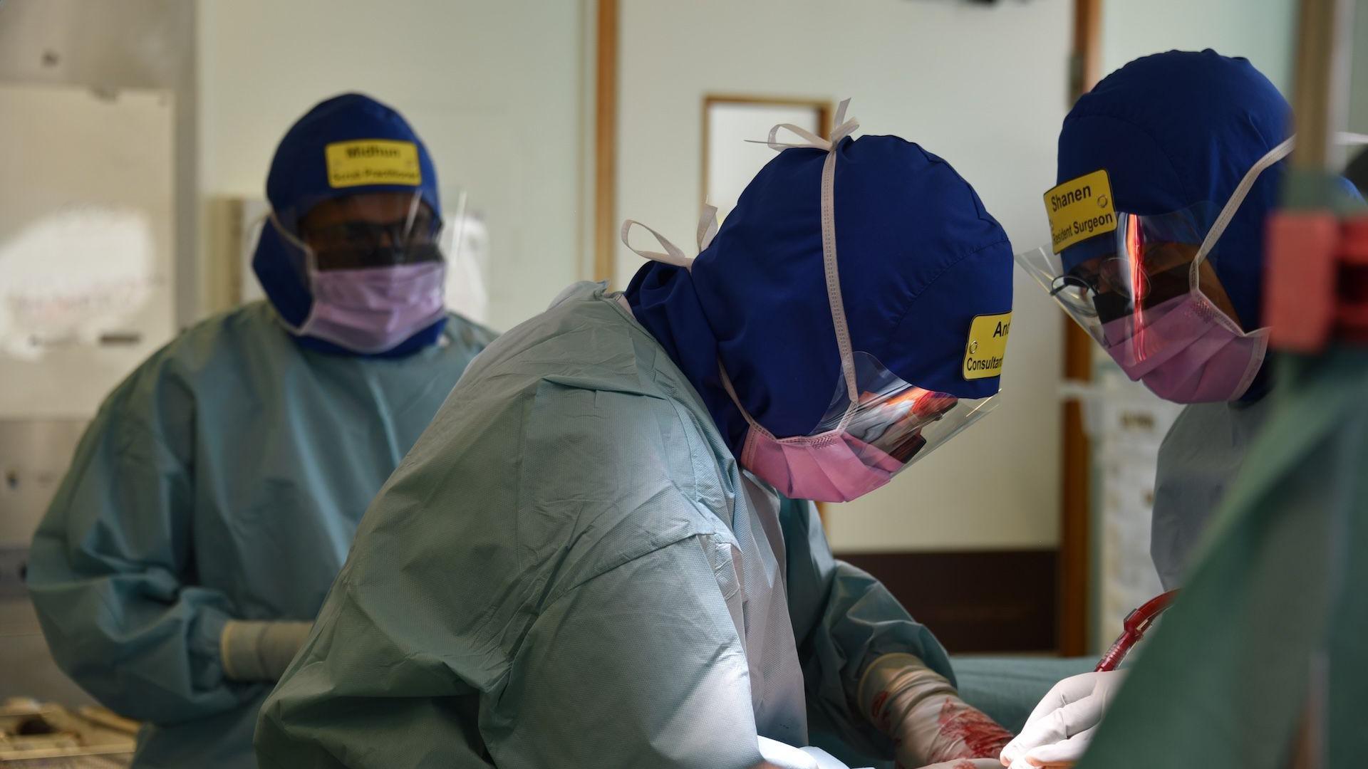 A group of people wearing medical scrubs and hats, with yellow name badges on. They're looking over a table as they are performing surgery.